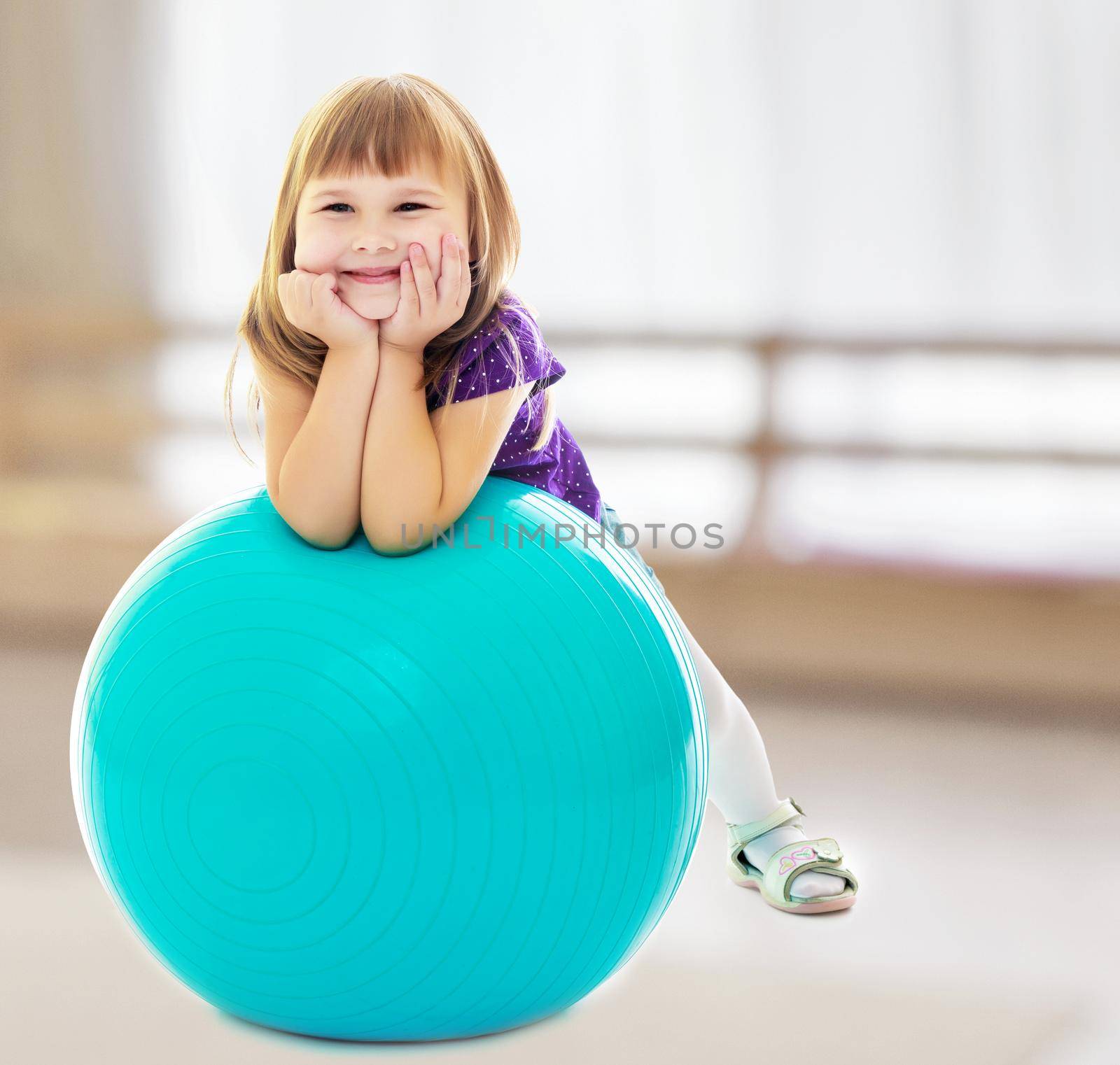 In a room with a large semi-circular window. About what dreams a little girl , put his hands on big blue fitness ball.