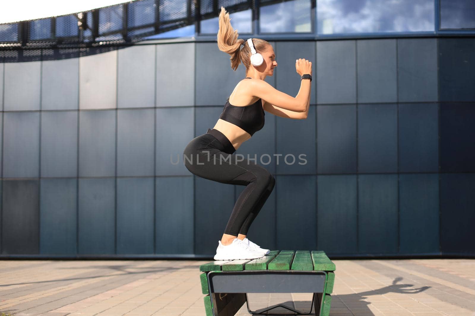 Young fit active woman in earphones doing jumps on a bench on city street