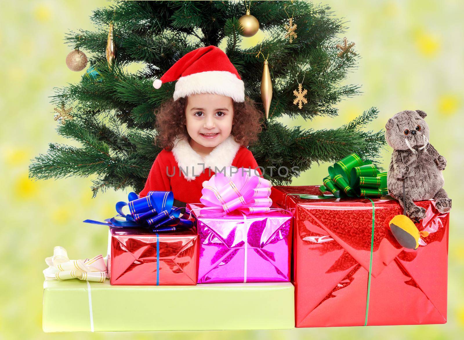 The beautiful, curly little girl in a suit and hat of Santa Claus sits near Christmas tree with bunch of gifts.Bright,floral yellow-green blurred background.