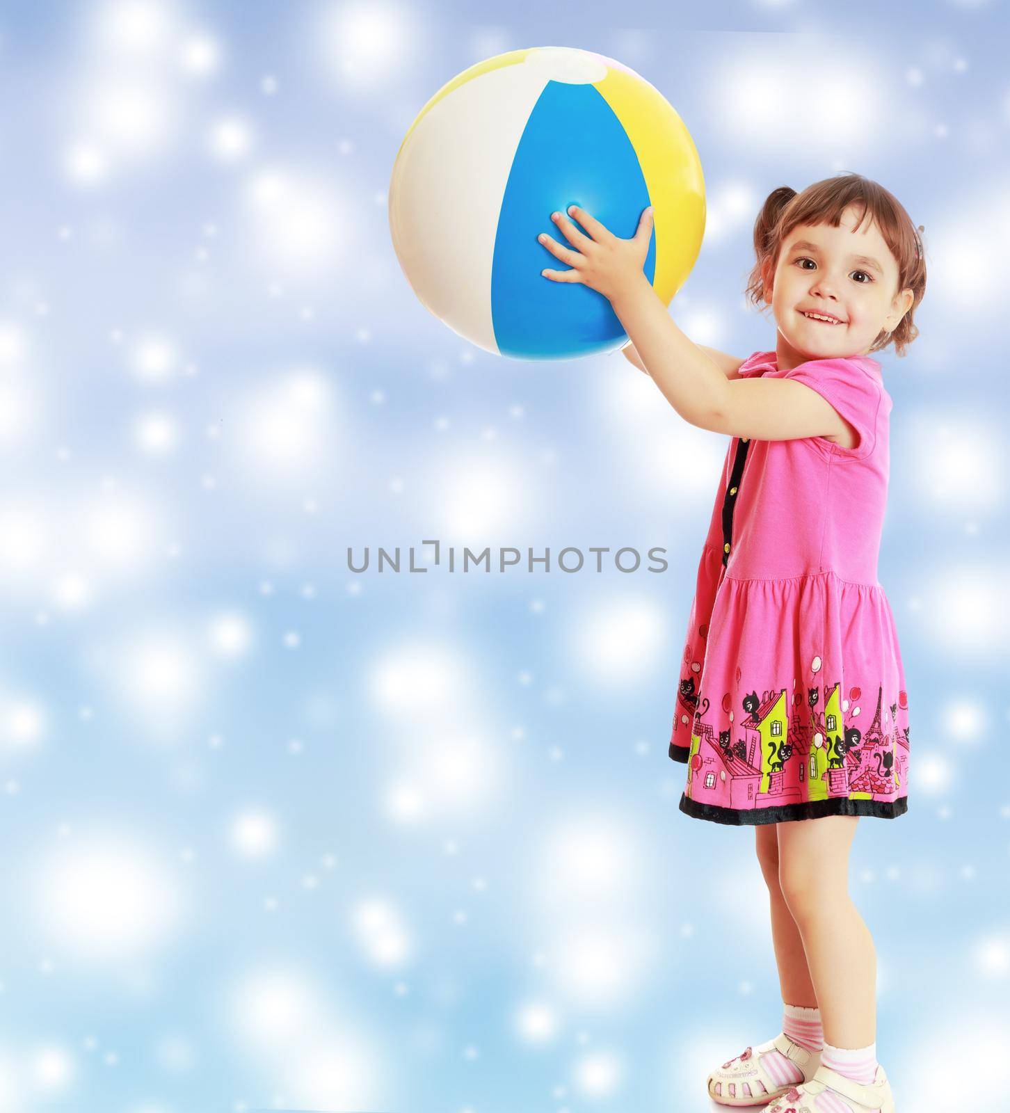 Happy little girl with pigtails on the head , in a pink dress. The girl lifted a large, inflatable striped ball.On new year or Christmas blue background with white big stars.