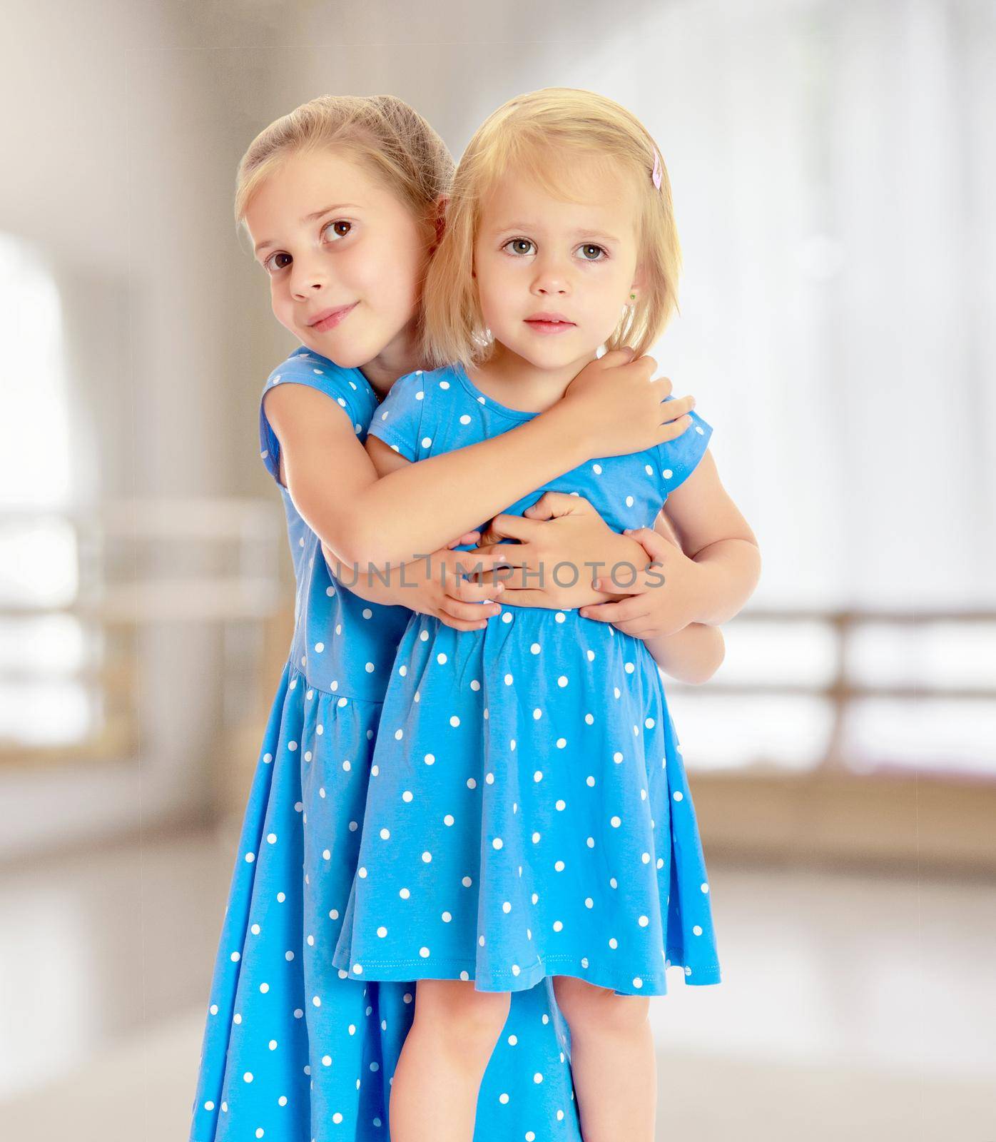 Two charming little girls, sisters , in identical blue dresses with polka dots , cuddling.