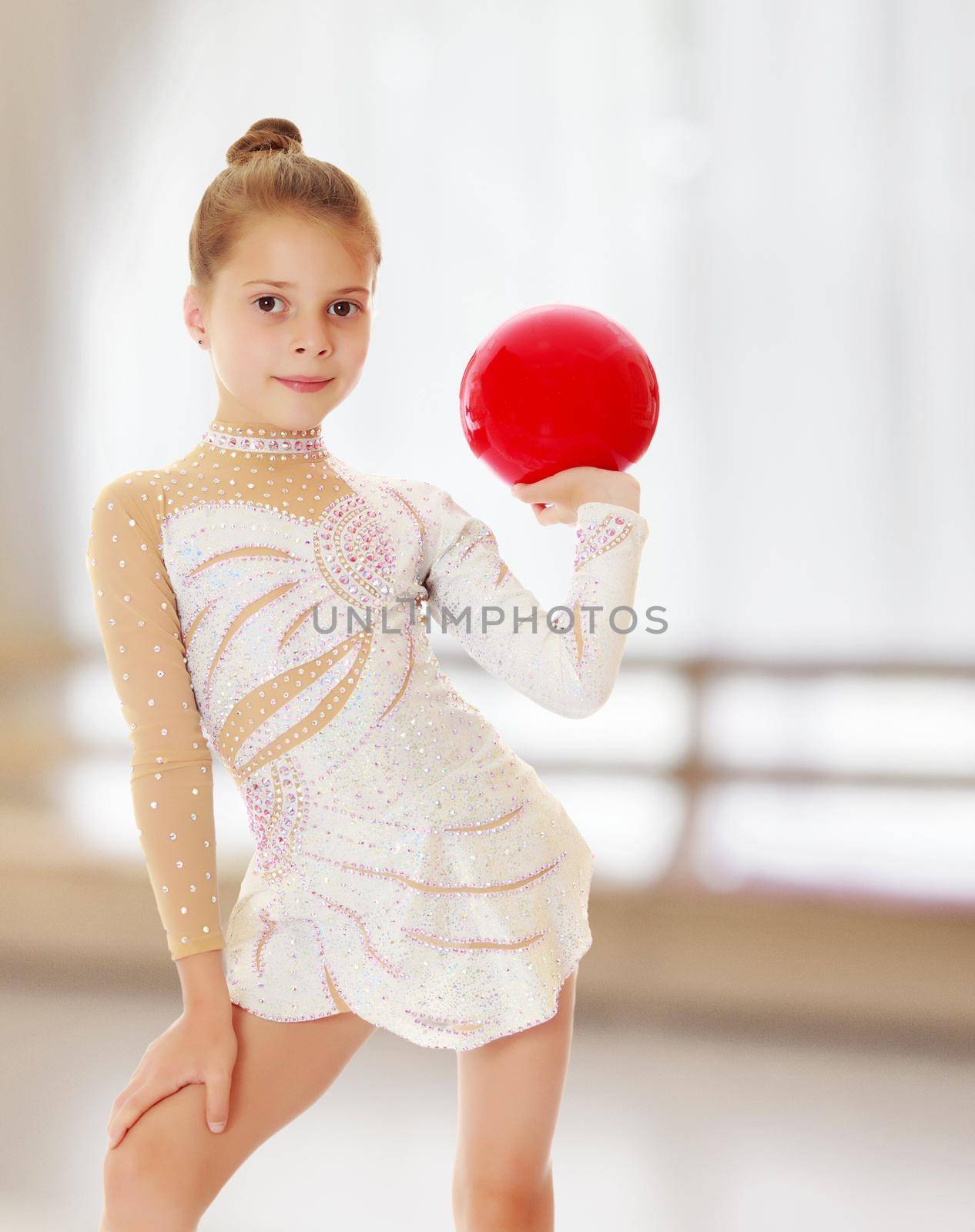 Beautiful little girl gymnast in elegant dress, posing with a red ball.On the background of the hall with large semi-circular window.