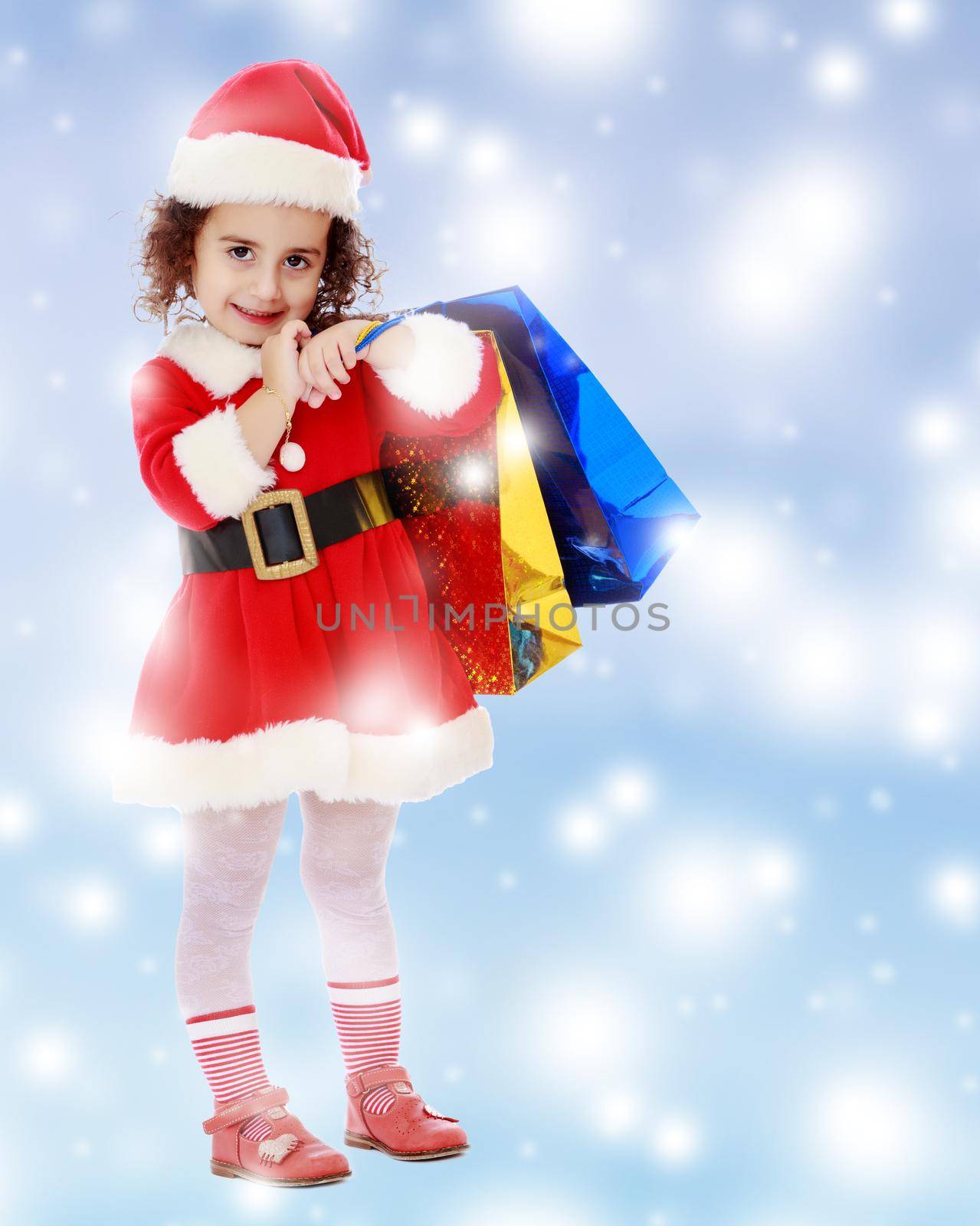 Caucasian little curly-haired girl in a coat and hat of Santa Claus carries on his shoulder a colorful shopping bags.Blue winter background with white snowflakes.