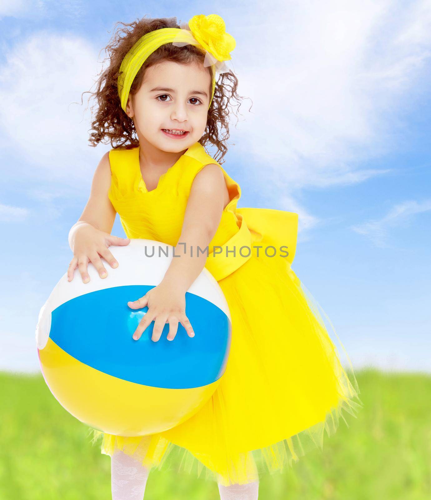 Close-up. Funny curly baby girl in a bright yellow dress and bow on her head playing with a ball.On the background of grass and sky.