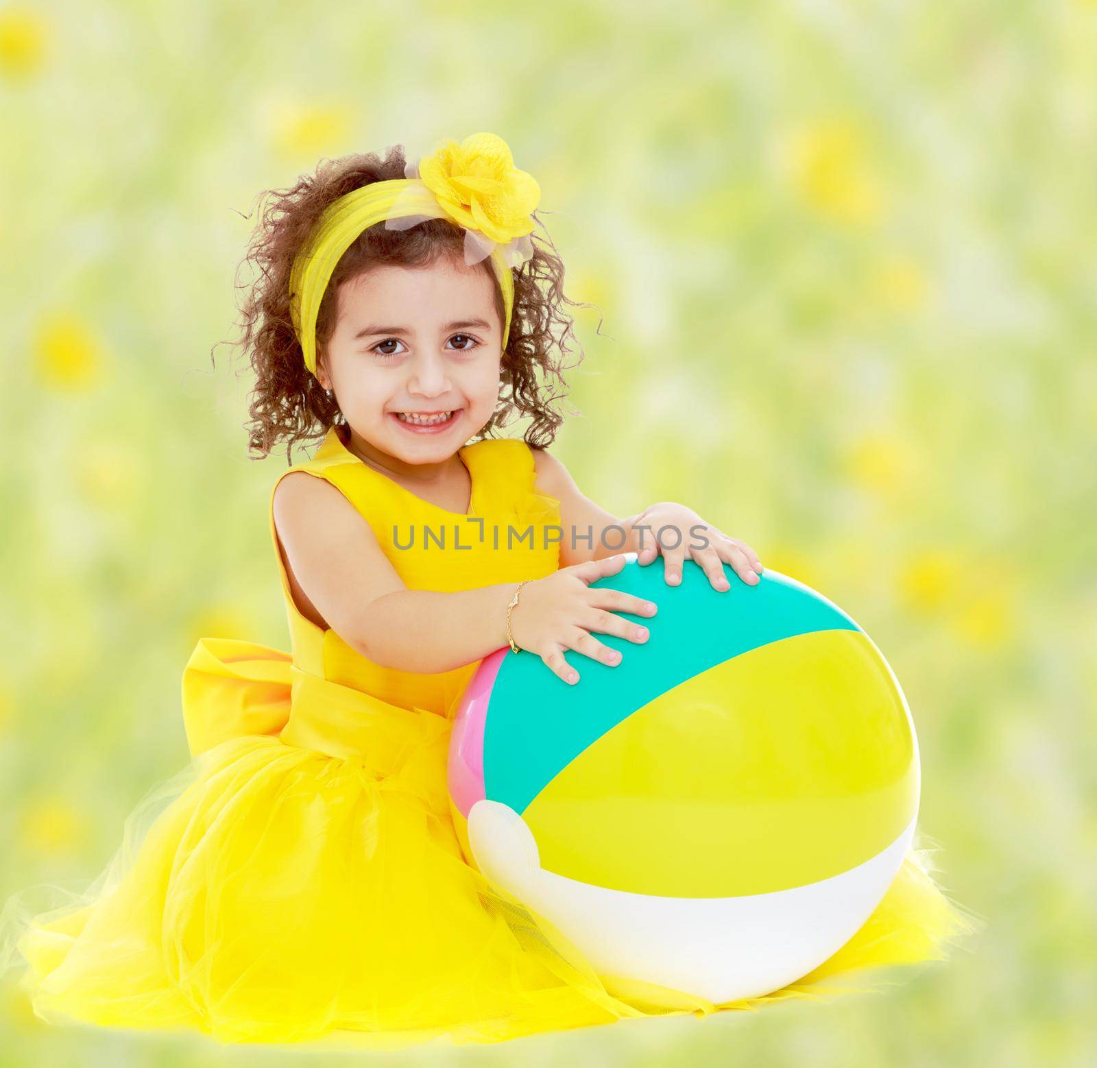 Joyful little girl in a yellow dress and bow on her head sitting on the floor. Girl hands hugging the ball.Bright,floral yellow-green blurred background.
