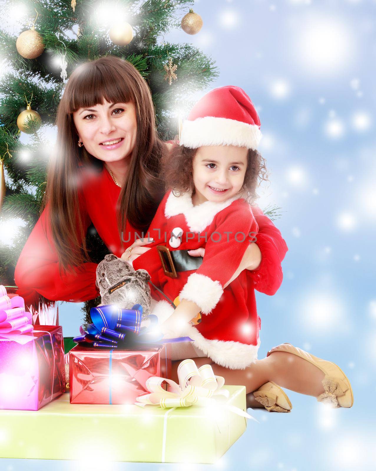 Happy mother and daughter near a Christmas tree surrounded by heaps of gifts.Blue winter background with white snowflakes.