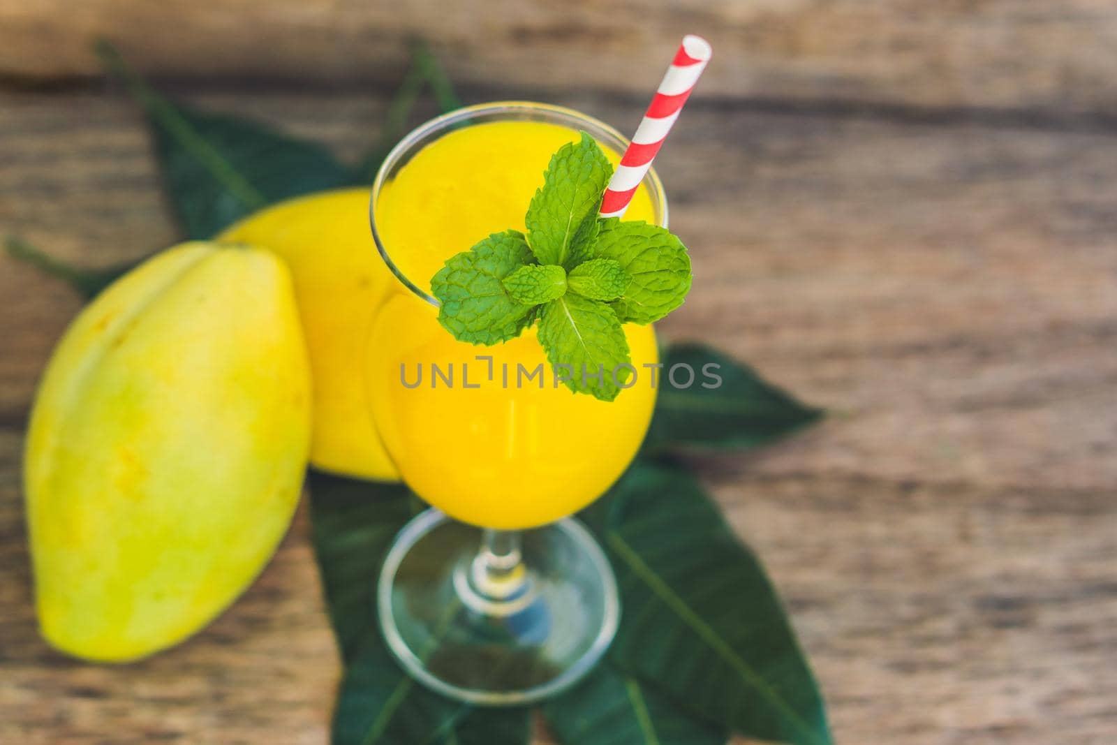 Juicy smoothie from mango in glass with striped red straw and with a mint leaf on old wooden background. Healthy life concept, copy space.