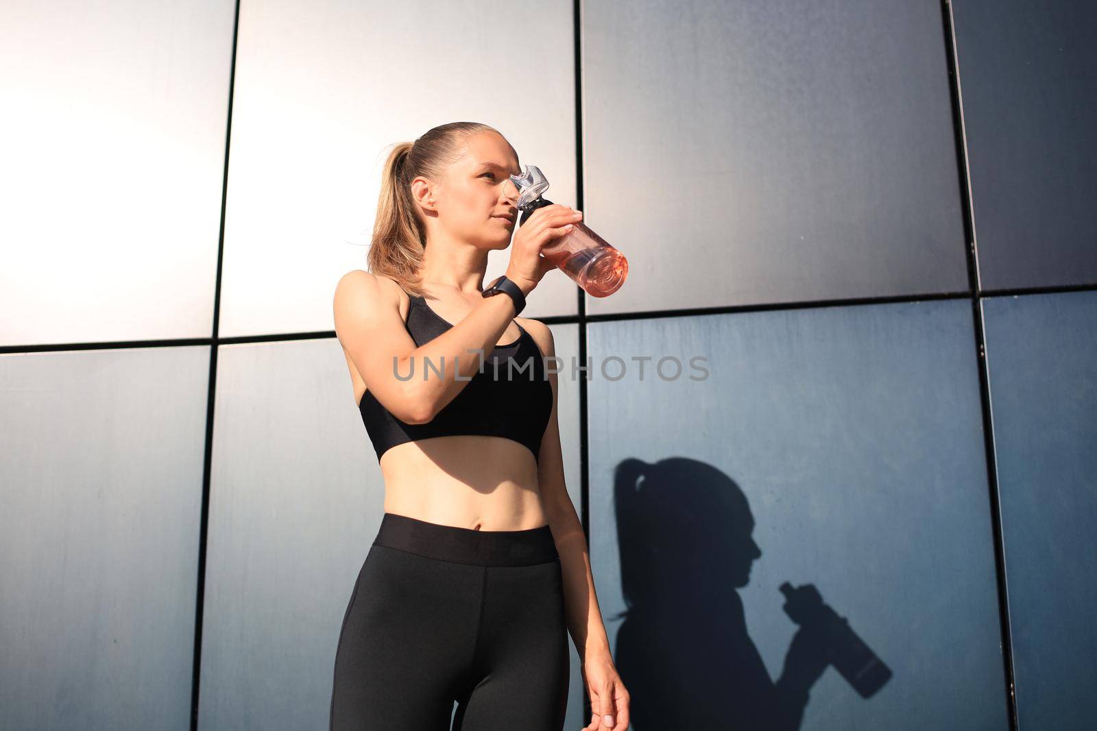Close up of a young attractive tired sportswoman outdoors, drinking water from a bottle