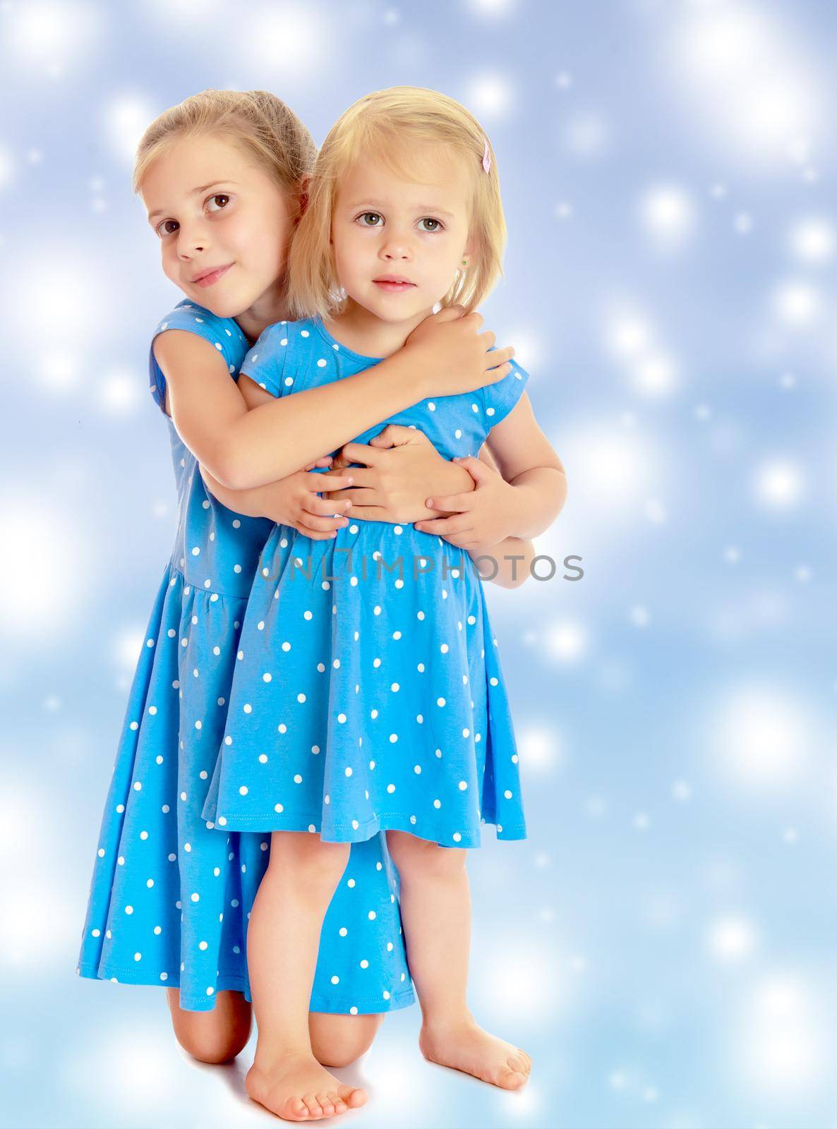 Two charming little girls, sisters , in identical blue dresses with polka dots , cuddling.On a blue background with large, white, Christmas or new year's snowflakes.