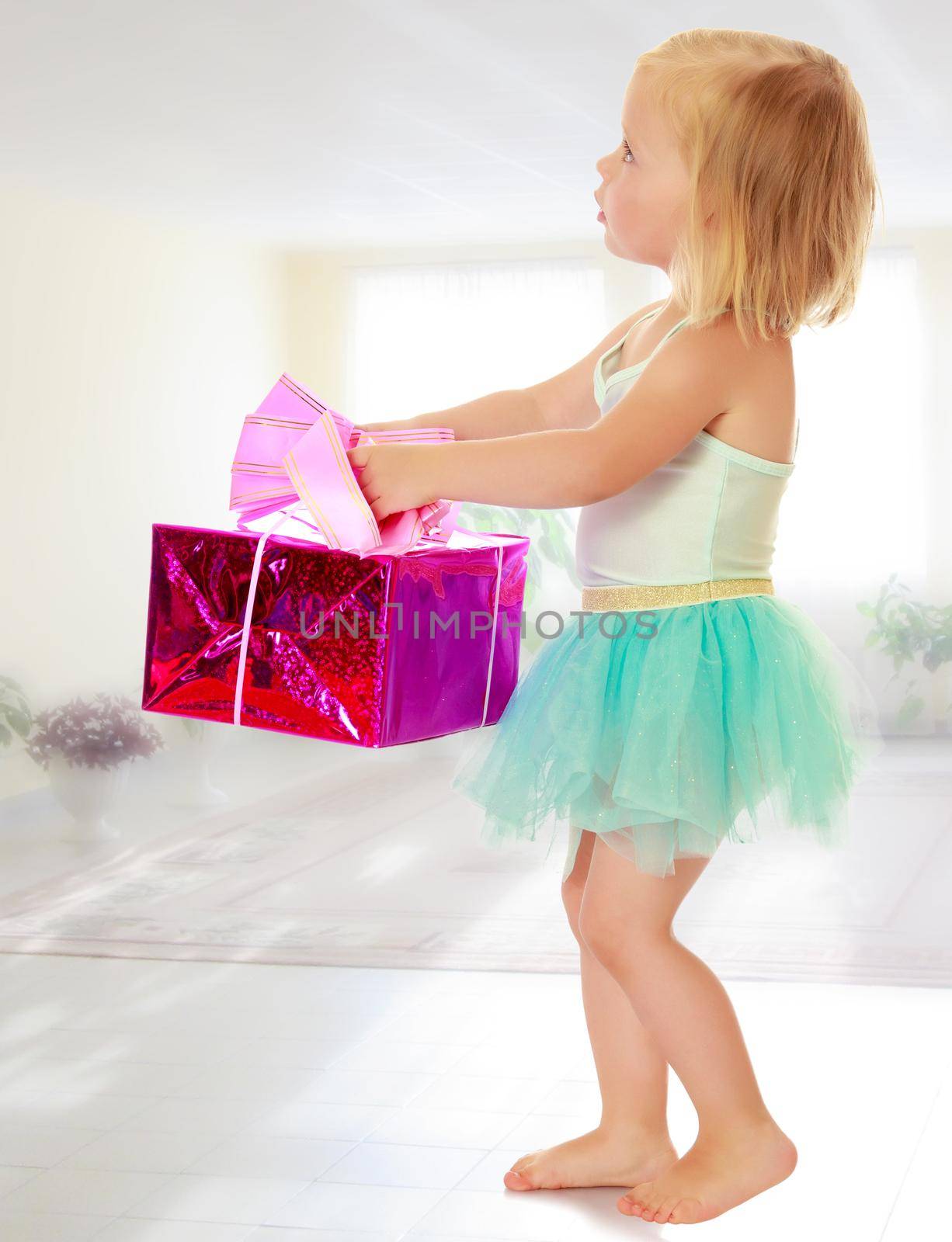 Cute little girl in a ballerina costume, carrying on his outstretched hands a gift box.On the background of the school hall with large Windows.