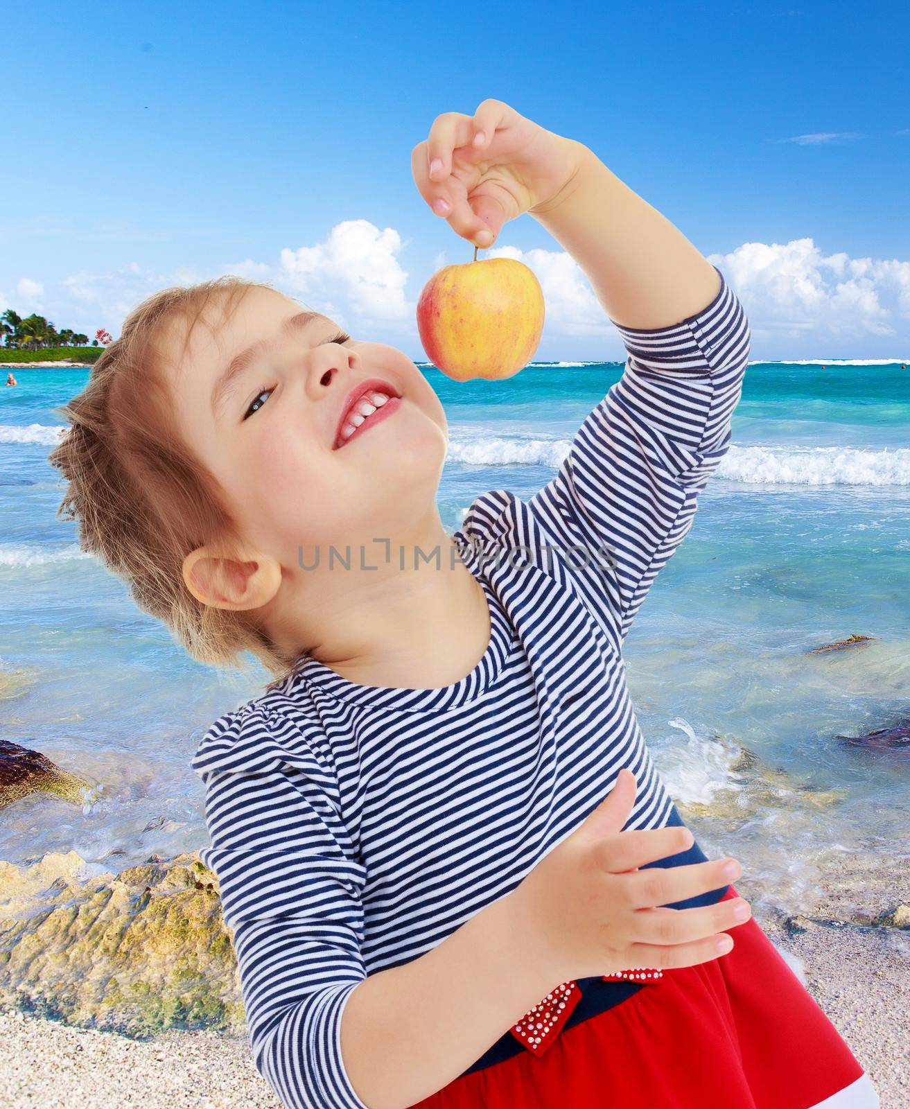 Nice little blonde girl with a red bow on the head holding the tail in front of the face of a delicious Apple . close-up.On the background of white, sandy beaches, blue sky and clouds.