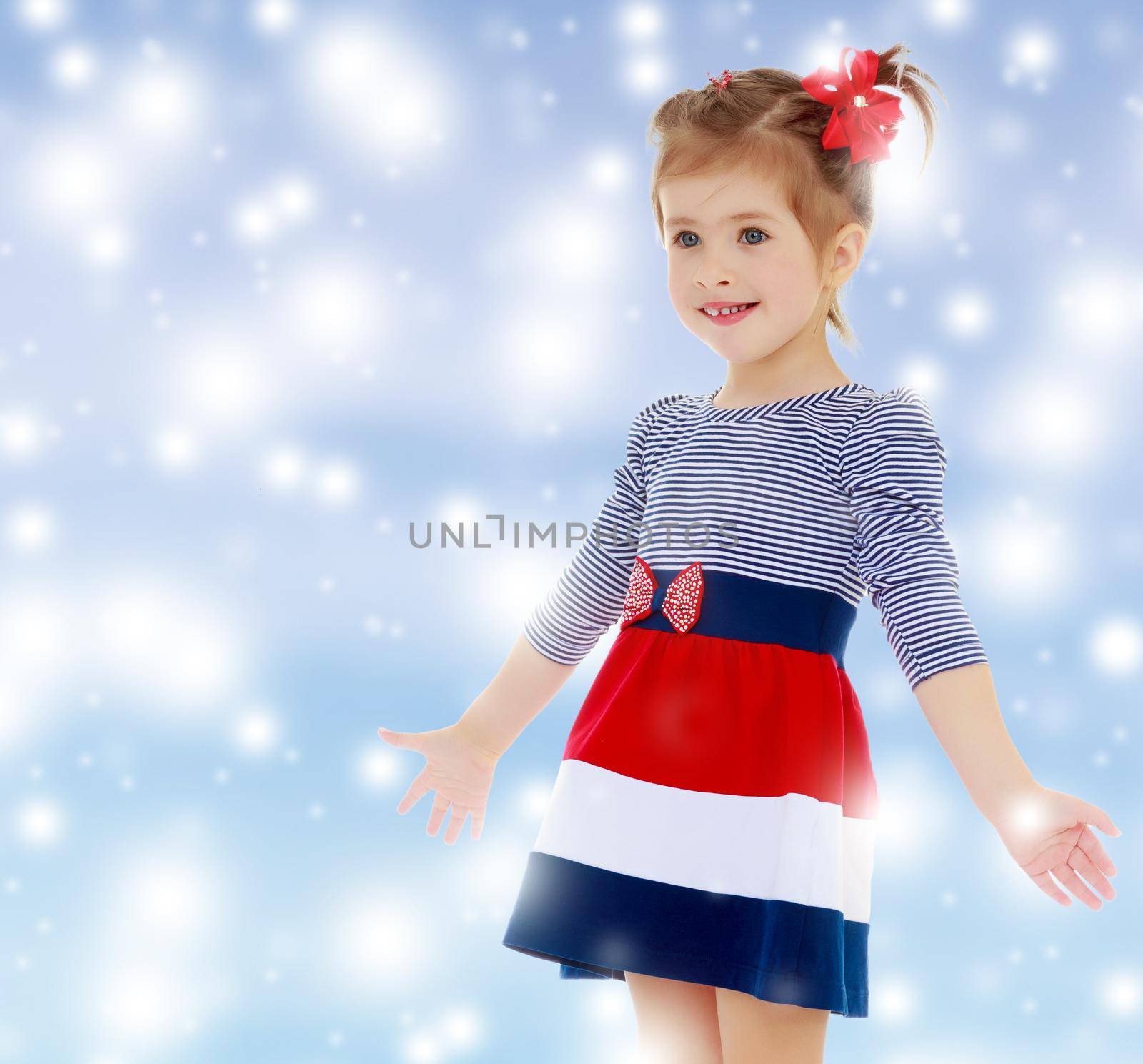 Little Caucasian blond girl with a red bow on her head, dressed in striped dress, she shrugs. Close-up.Gentle blue Christmas background with white snowflakes abstract.