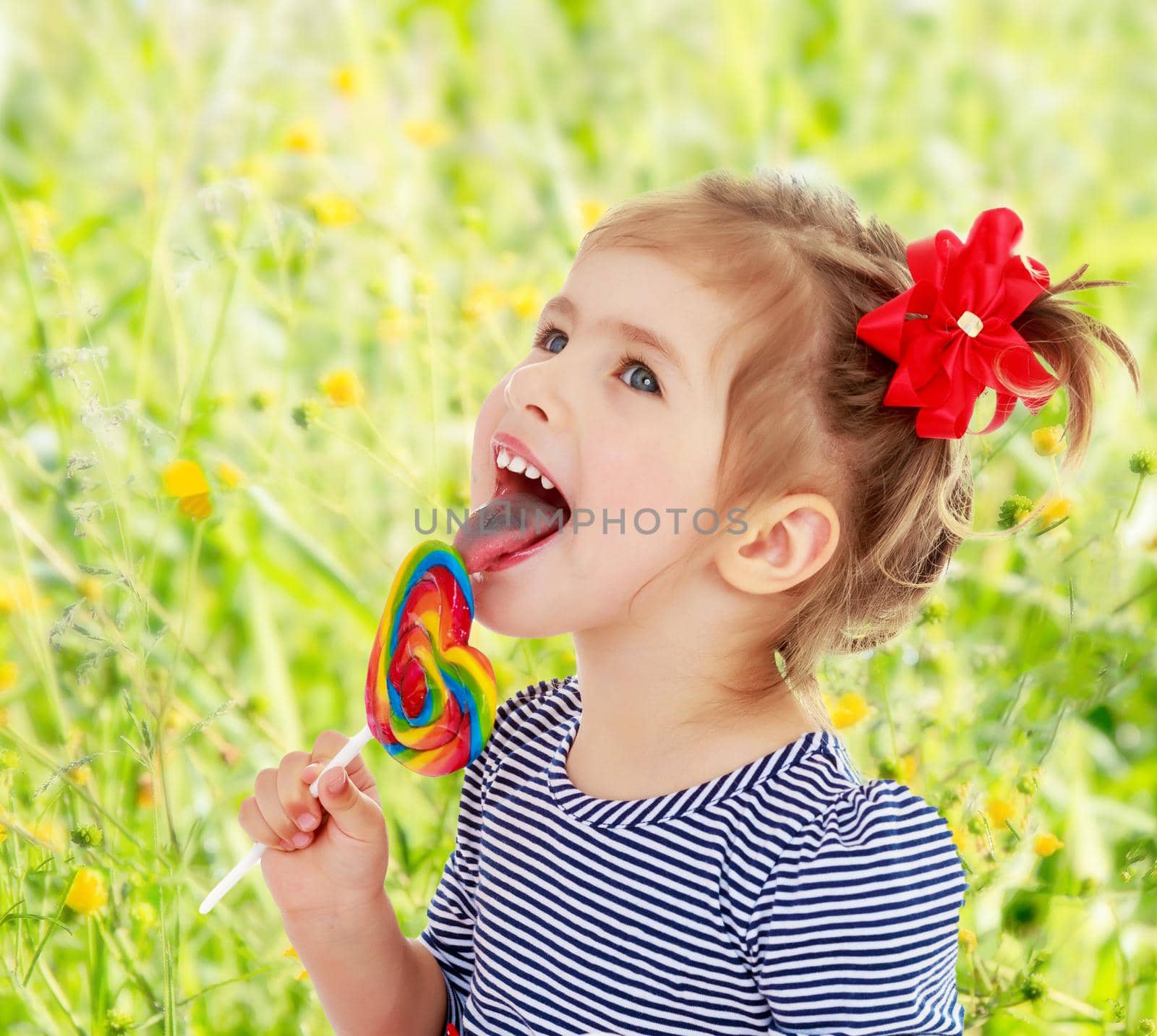 Cute little blonde girl with a red bow on her head, with pleasure licking colorful candy on a stick. Visible language which was painted in a candy color. Close-up.On blurred background of green grass