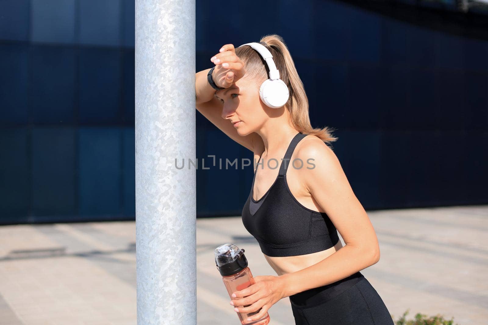 Beautiful female runner is standing outdoors holding water bottle. Fitness woman takes a break after running workout