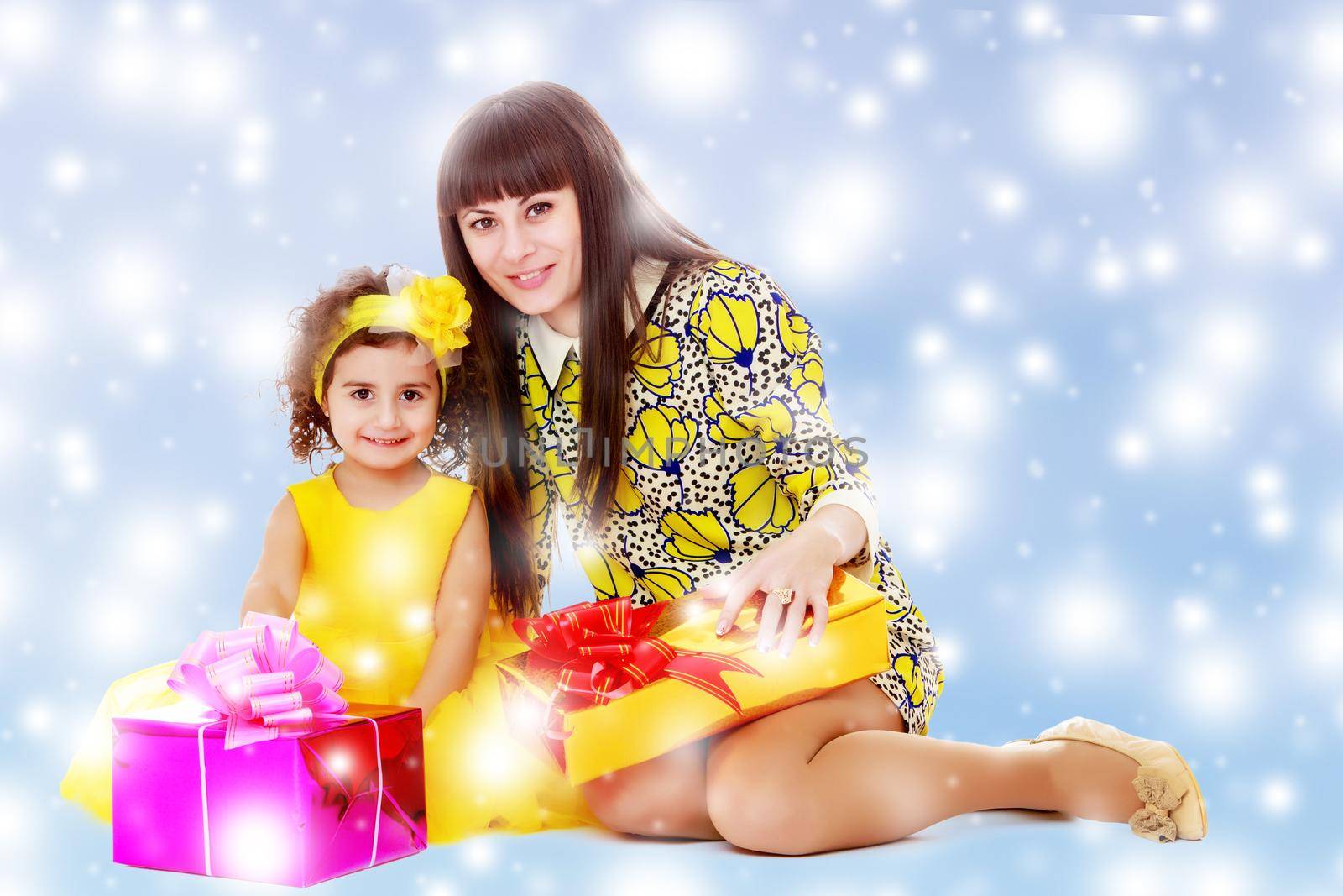 Beautiful young mother and her adorable little daughter , sitting surrounded by Christmas gifts.Blue winter background with white snowflakes.