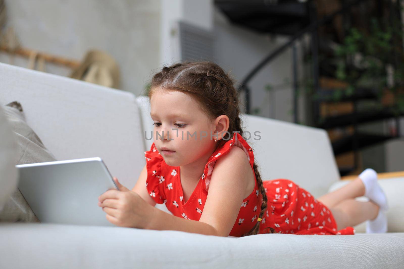 Smiling little girl lying on sofa playing online games, web surfing information, using funny applications on tablet. by tsyhun