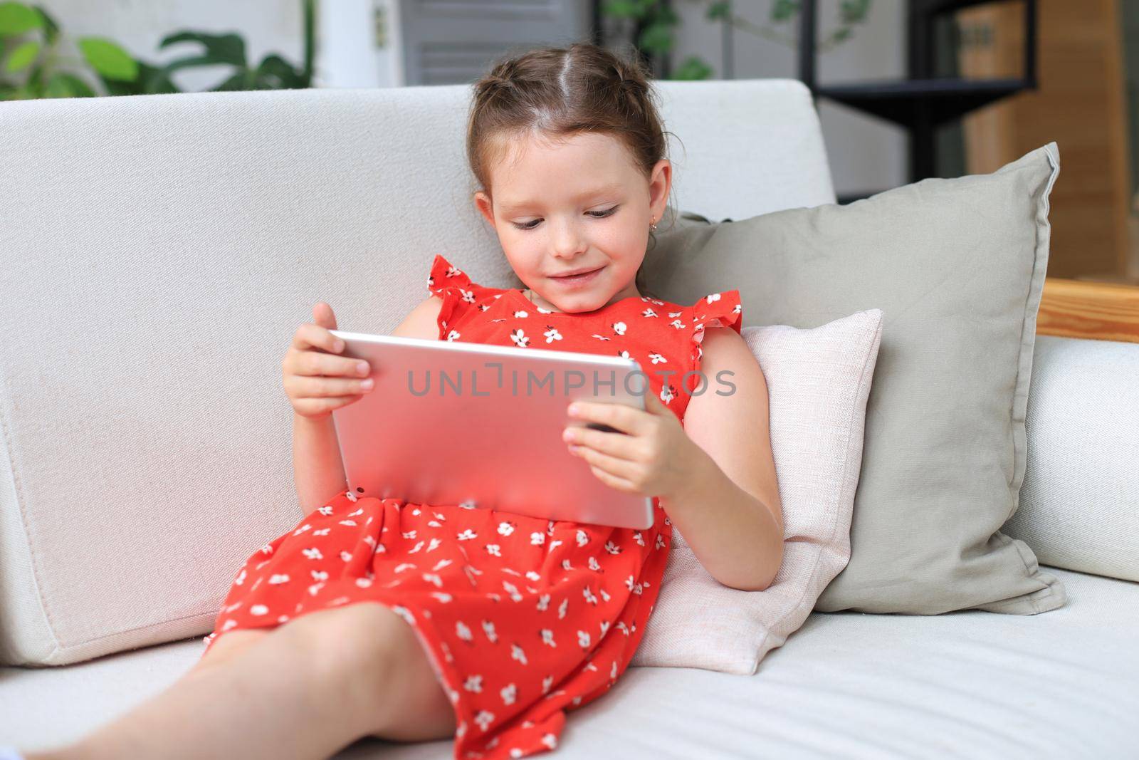 Smiling little girl lying on sofa playing online games, web surfing information, using funny applications on tablet