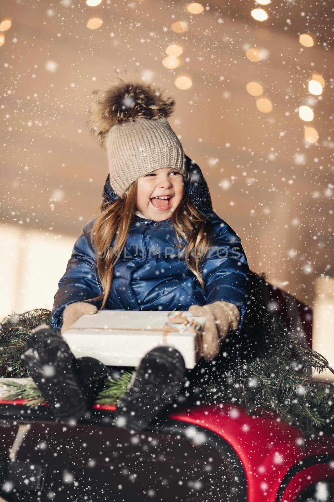 Little pretty girl sith wavy hair sits of the sofa with a box with a present and rejoices by StudioLucky