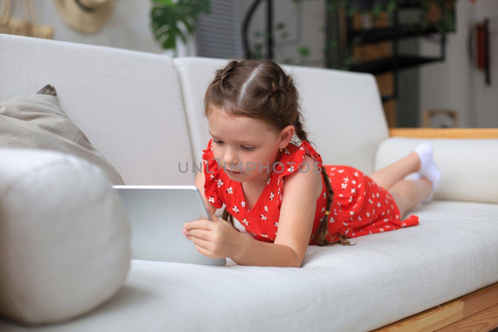 Smiling little girl lying on sofa playing online games, web surfing information, using funny applications on tablet
