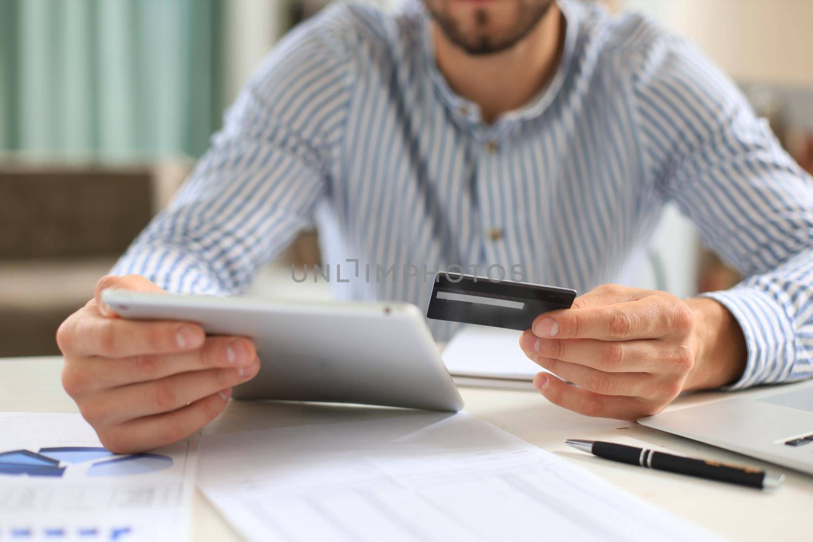 Young man sitting in office and pays by credit card with his tablet