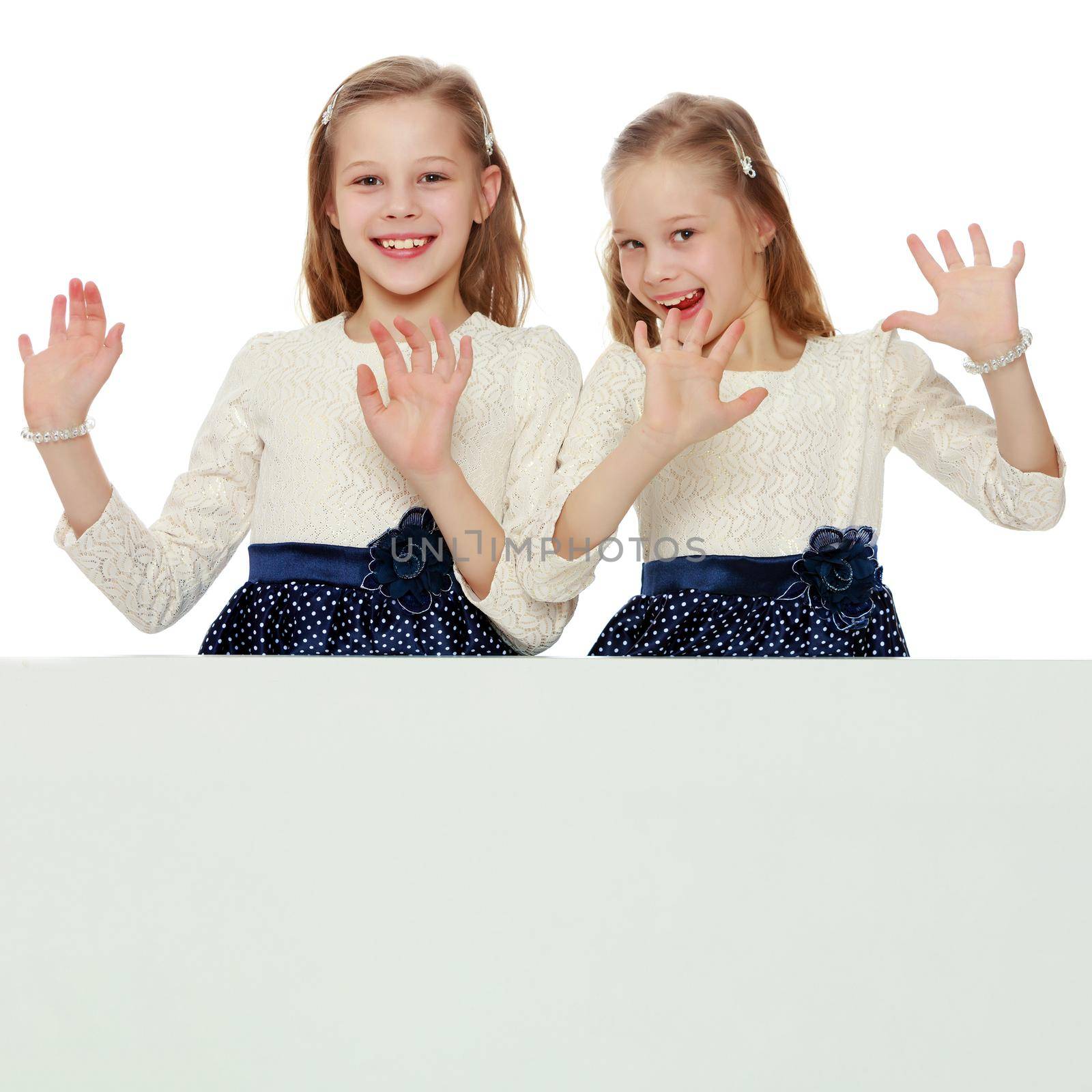 Two cute little girls look out from behind a white advertising banner. On the banner you can make any advertising inscription or put a picture or logo. It is isolated on a white background.