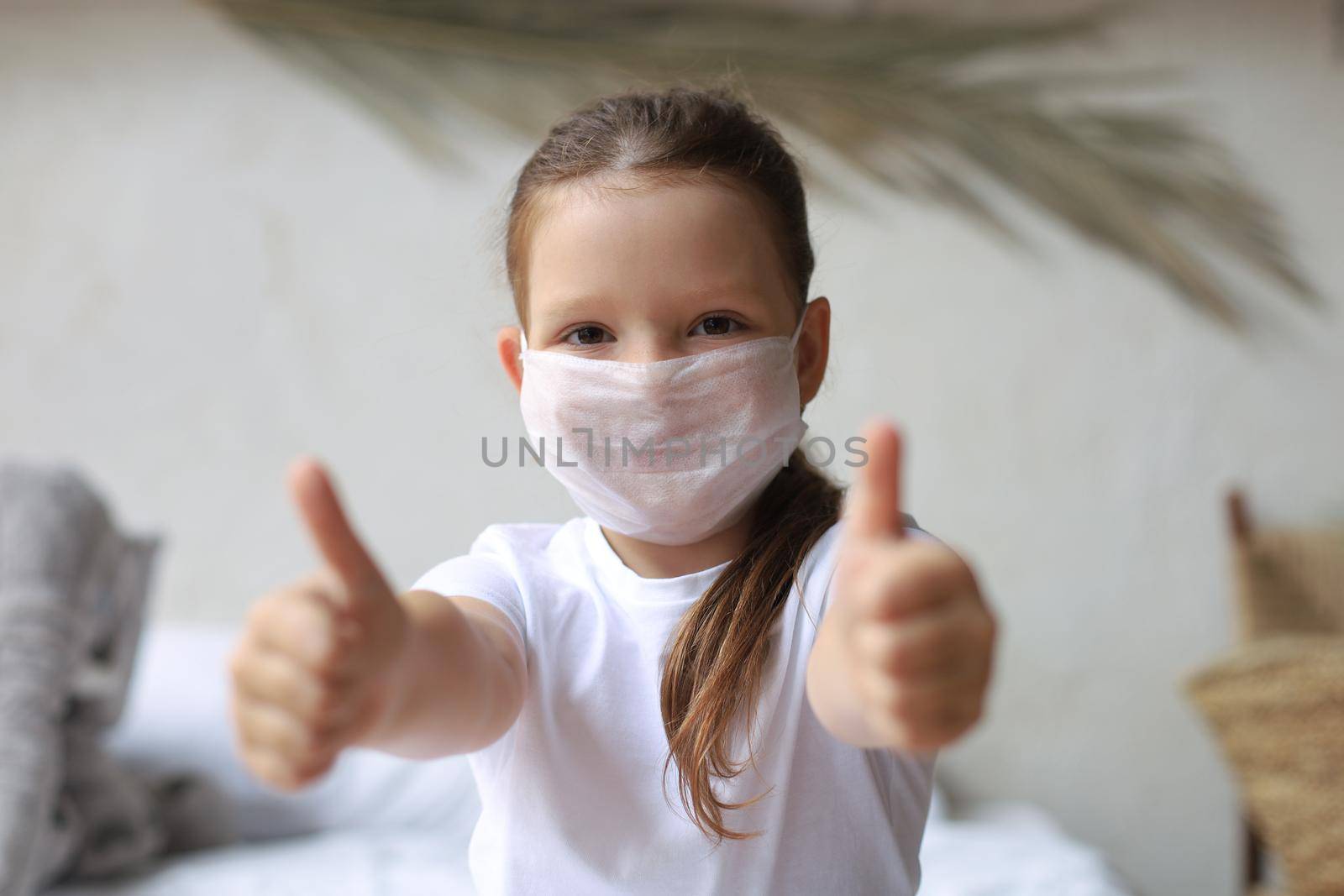 Little girl in a medical mask showing thumb's up gesture. Protection against coronavirus