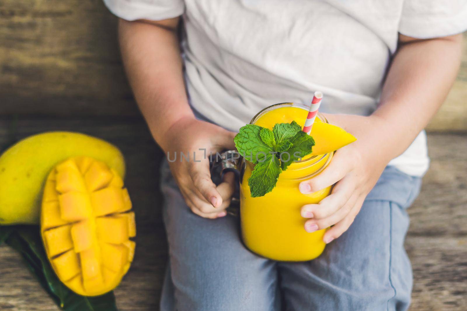 Boy drinking juicy smoothie from mango in glass mason jar with striped red straw on old wooden background. Healthy life concept, copy space by galitskaya