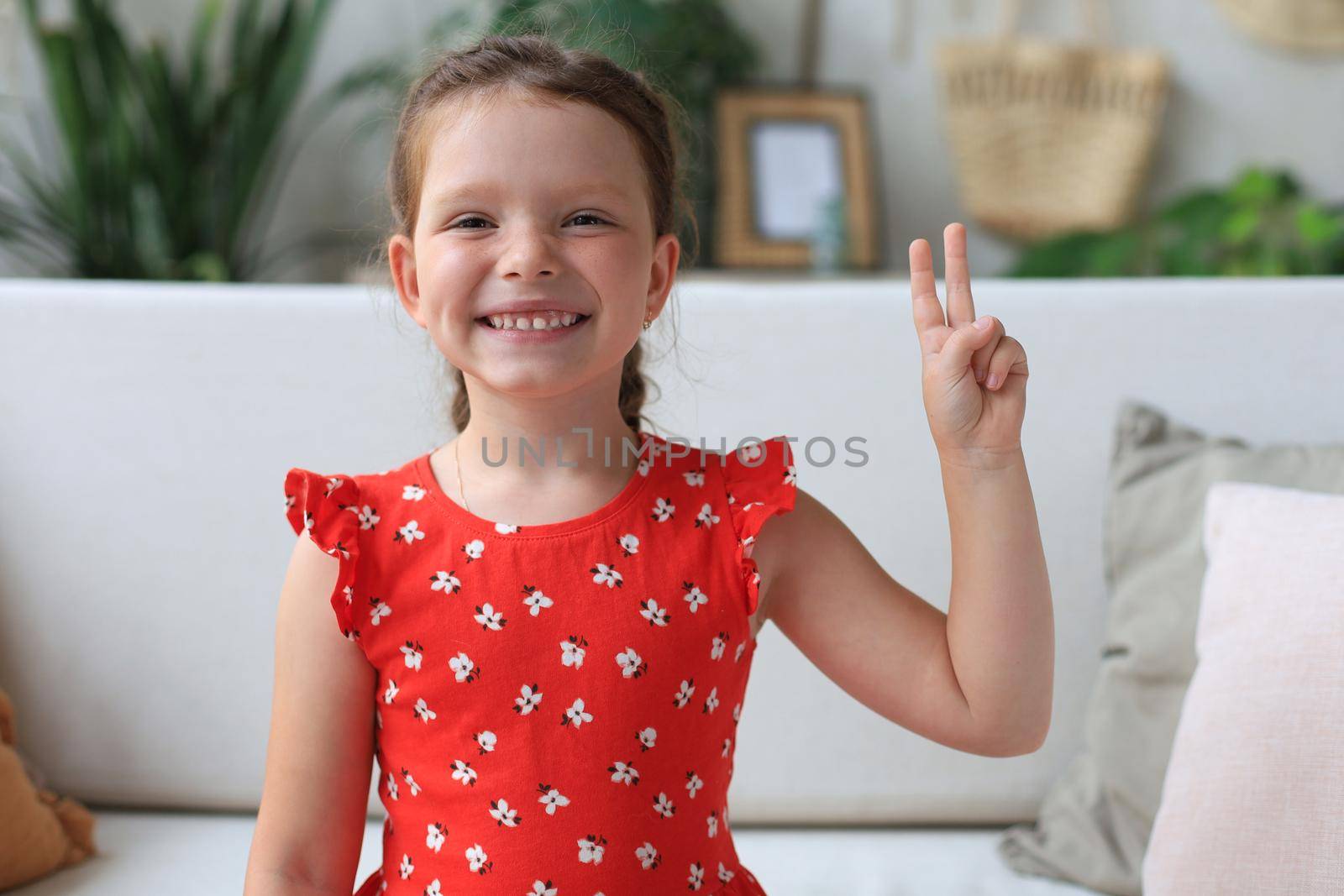 Little caucasian girl in red dress sitting on sofa, smiling with happy face doing victory sign