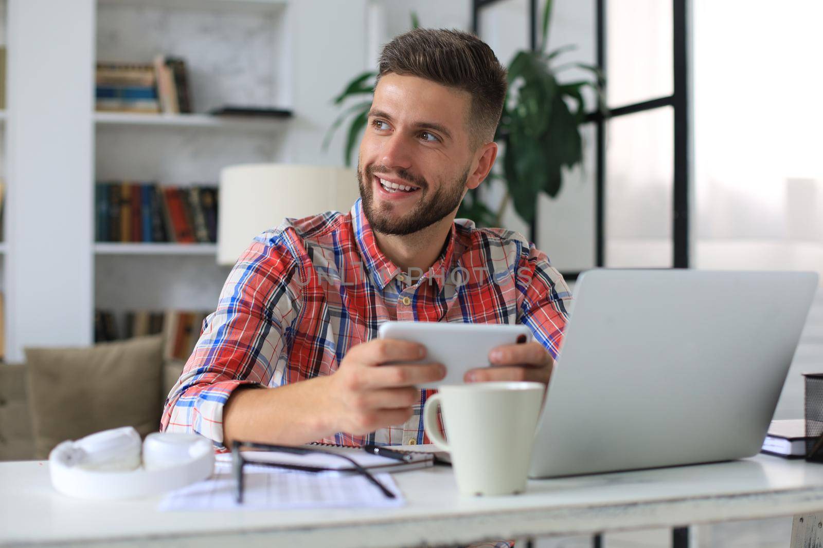 Attractive young man sitting at desk at home office and using mobile phone for cheking social nets