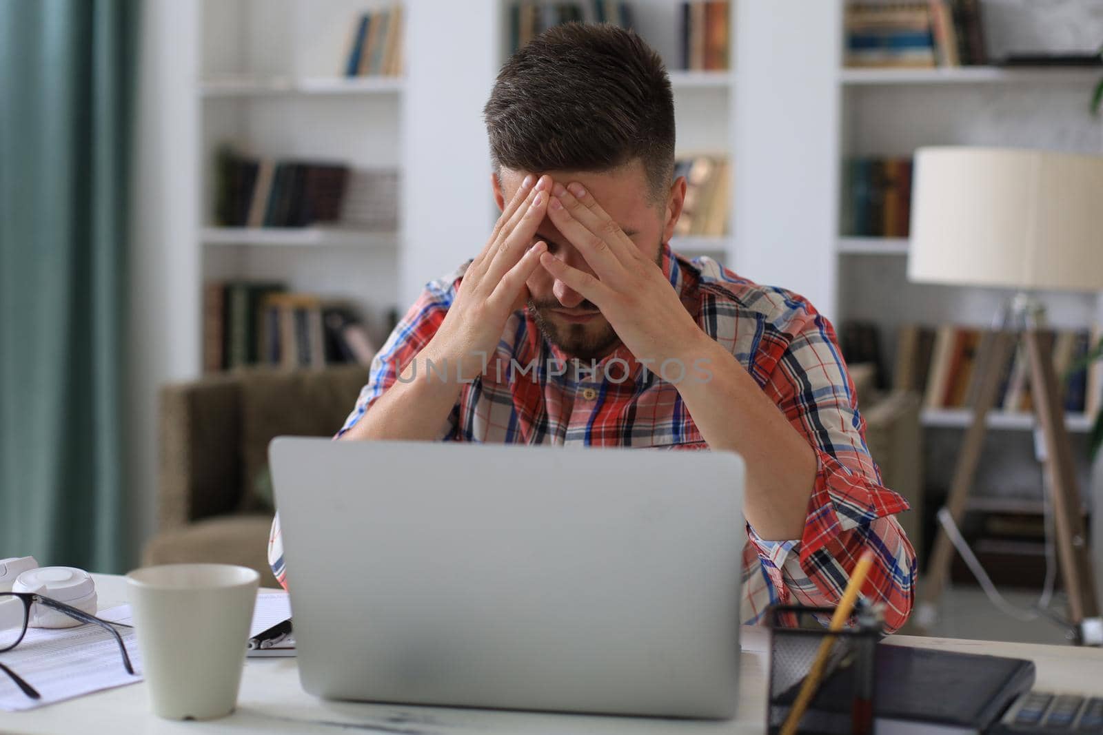 Unhappy frustrated young male holding head by hands sitting with laptop behind desk at home