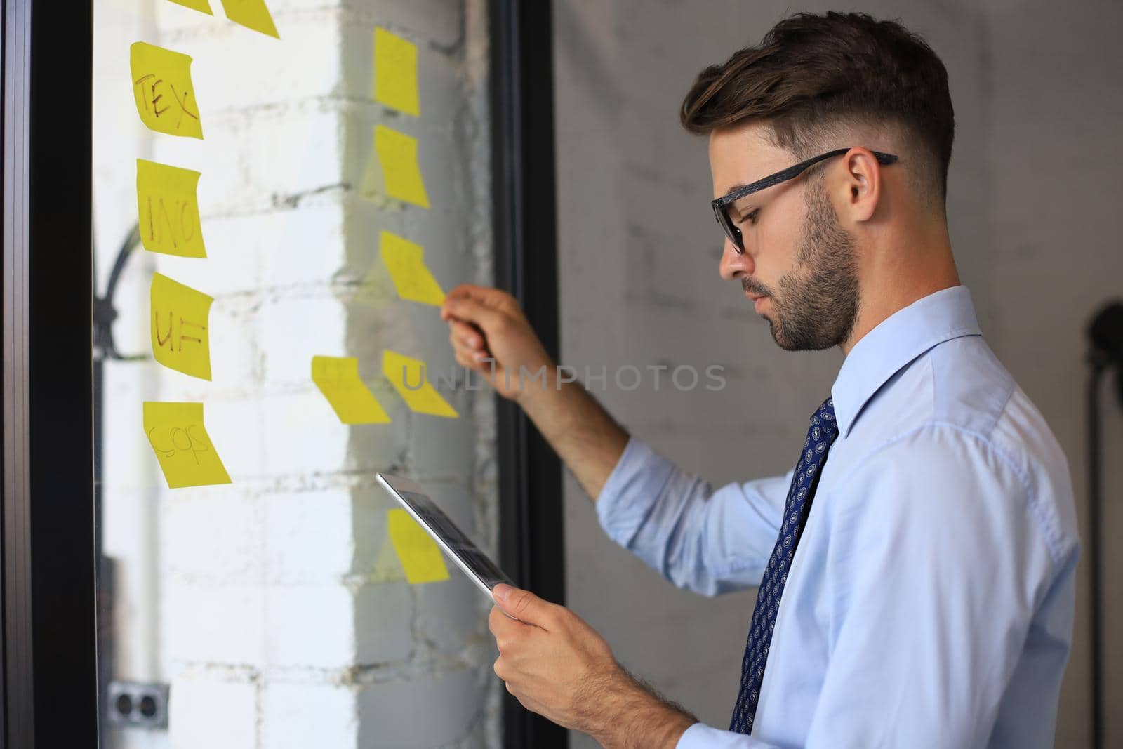 Young modern business man using adhesive notes while standing behind the glass wall in the office