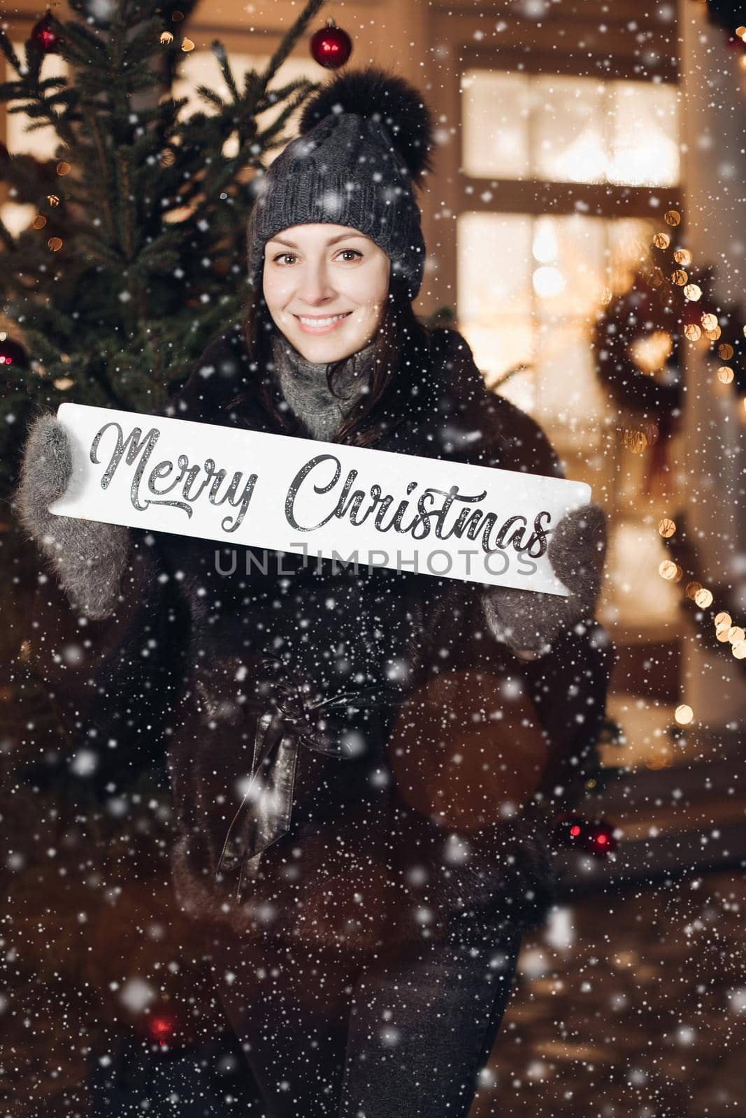 Attractive young woman in warm clothes stands under the snow and wishes everyone a merry Christmas by StudioLucky