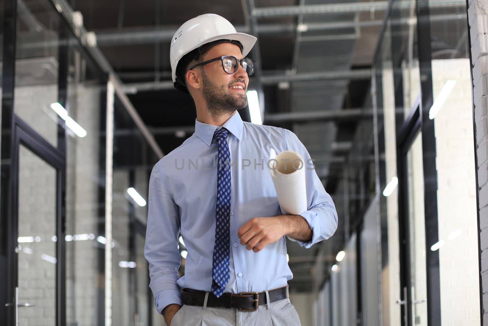 Confident young business man in hardhat holding blueprint and looking away while standing indoors