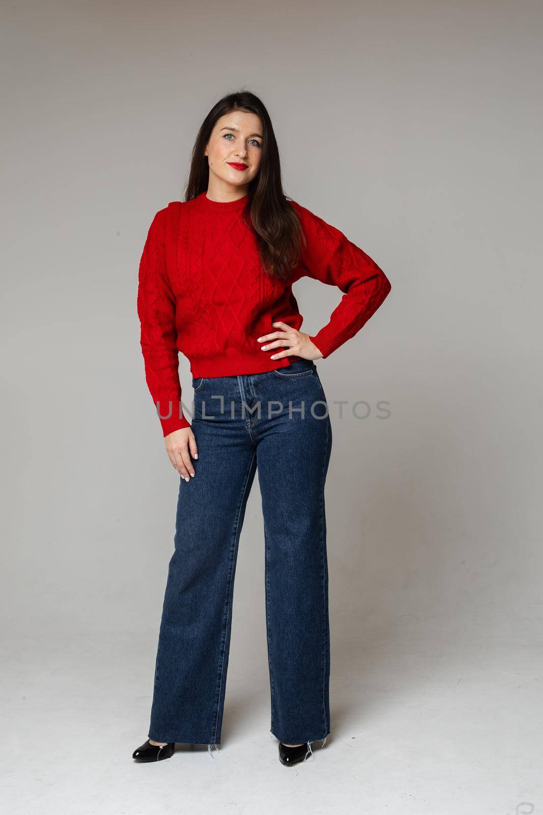 Studio portrait of beautiful young brunette woman in red knitted woolen sweater and stylish jeans and heels posing with hand at waist.