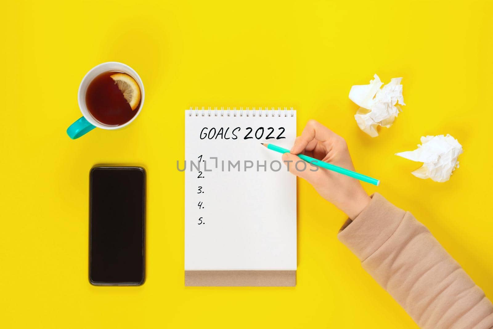 2022 goals banner with copy space. Notepad, pencil and cup of tea on a yellow background near crumpled sheets of paper and mobile phone. Copy space, Flat lay. Women hand write goals for 2020 year.