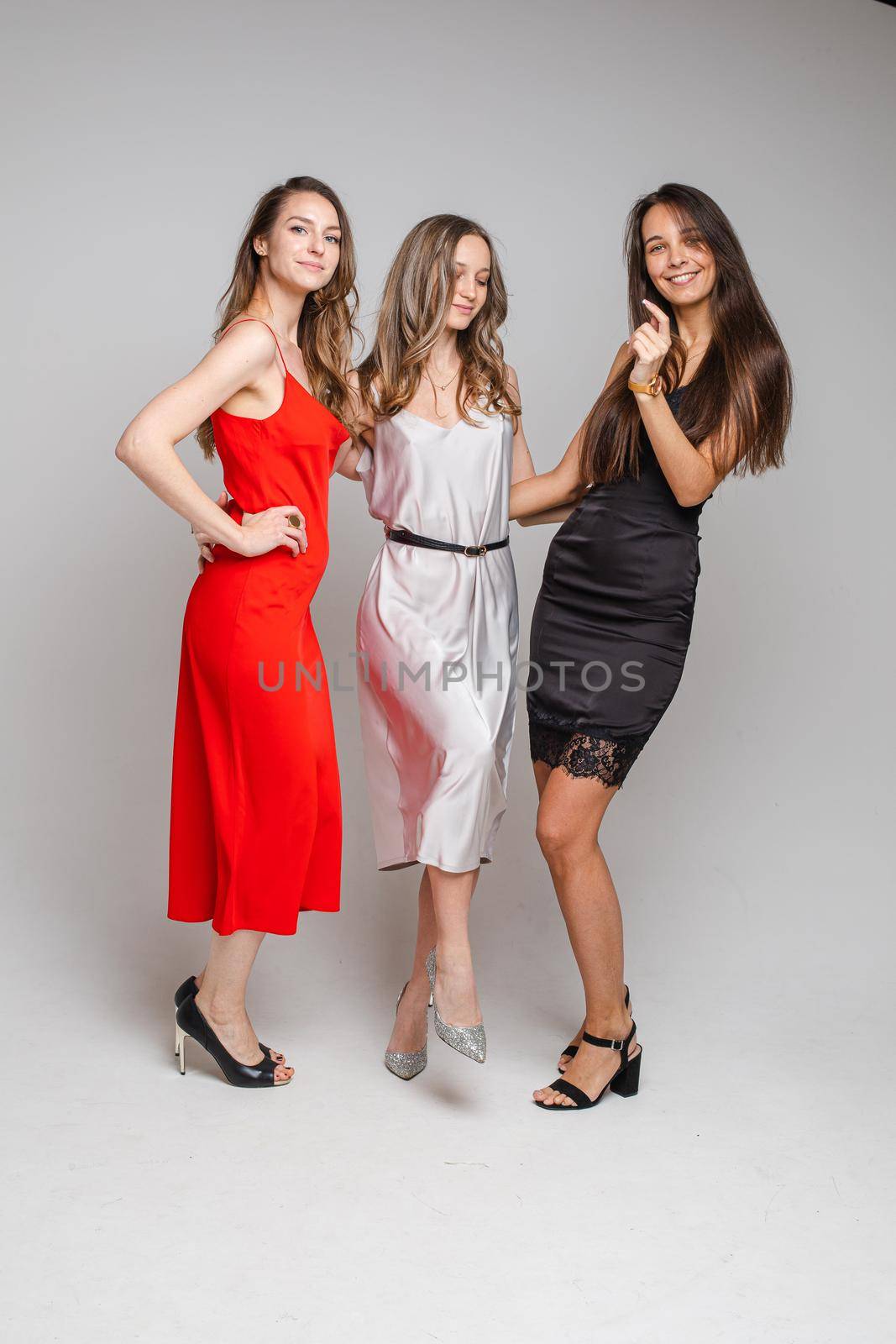 Charming positive young girls friends wearing stylish feminine evening dresses posing on gray studio background by StudioLucky
