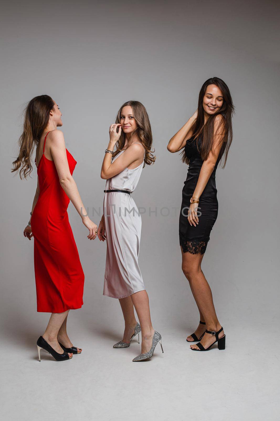 Full length portrait of gorgeous ladies in red, silver and black cocktail dresses and heels laughing all together on white background in studio.