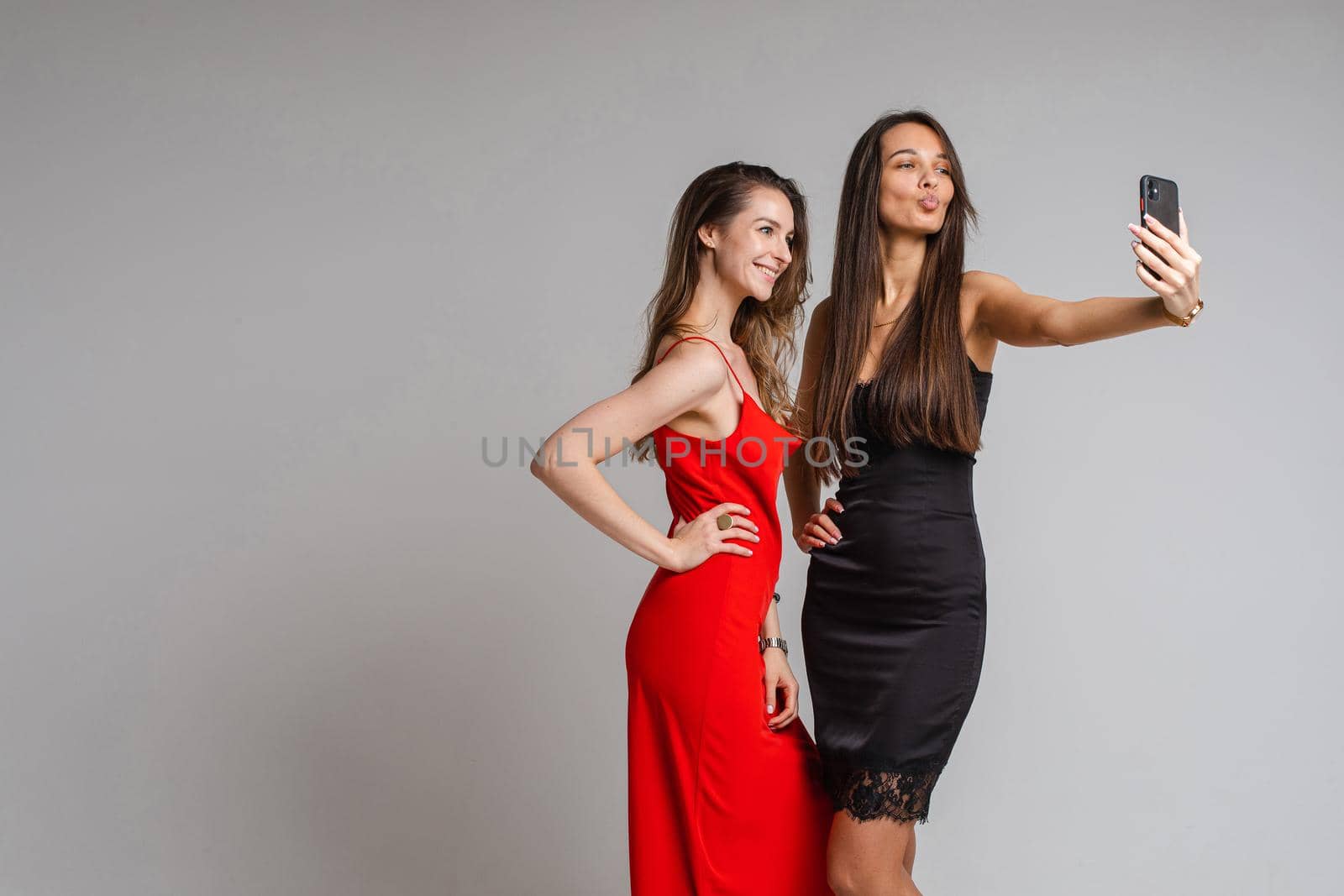 Happy beautiful two female friends posing in studio while making selfie on smartphone, isolated on grey background. Party concept