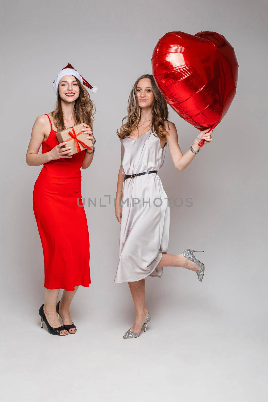 Happy two pretty women with heart-shaped balloon and gift box posing in studio, isolated on grey background. Party concept