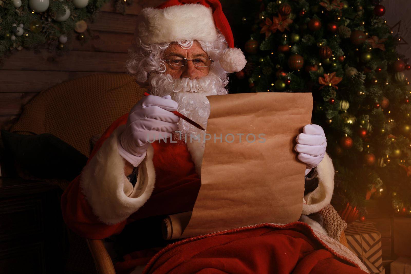 Portrait of happy Santa Claus sitting at his room at home near Christmas tree and reading Christmas letter or wish list