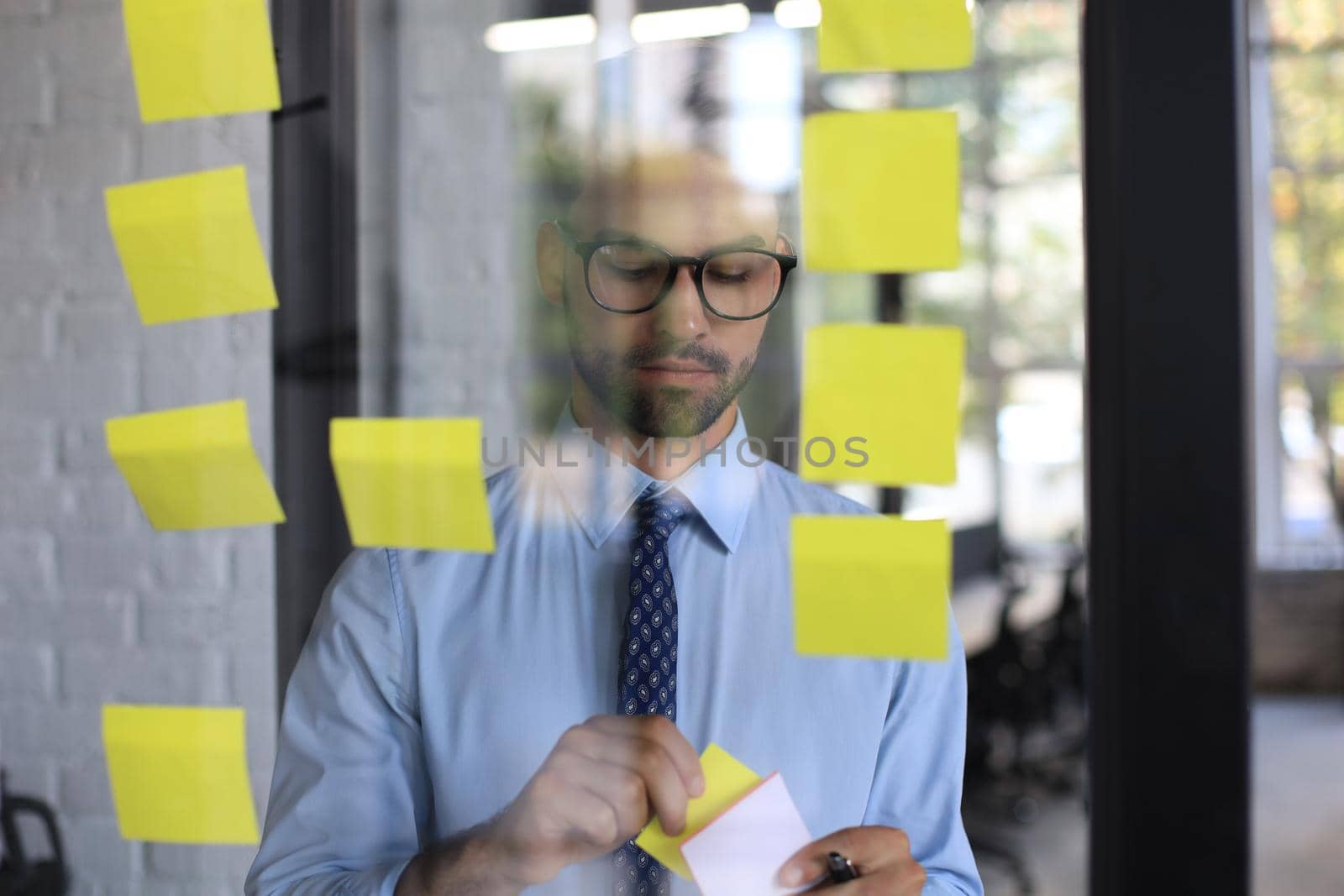 Young modern business man using adhesive notes while standing behind the glass wall in the office