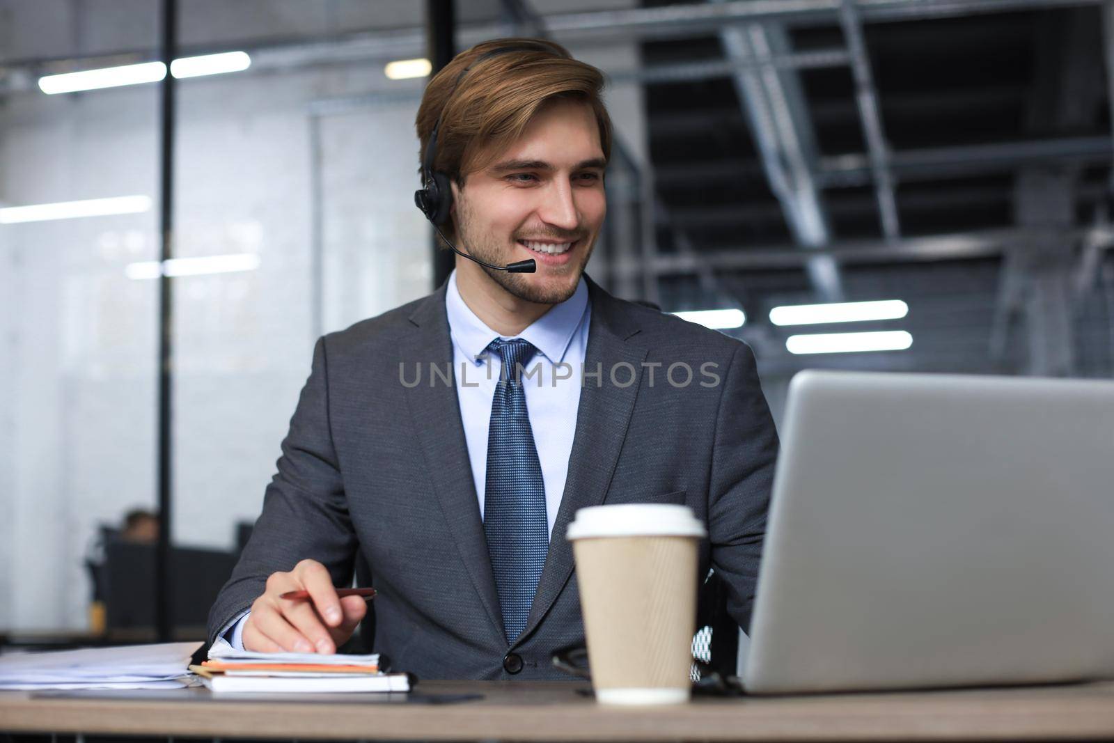 Smiling male call-center operator with headphones sitting at modern office, consulting online information in a laptop, looking up information in a file in order to be of assistance to the client