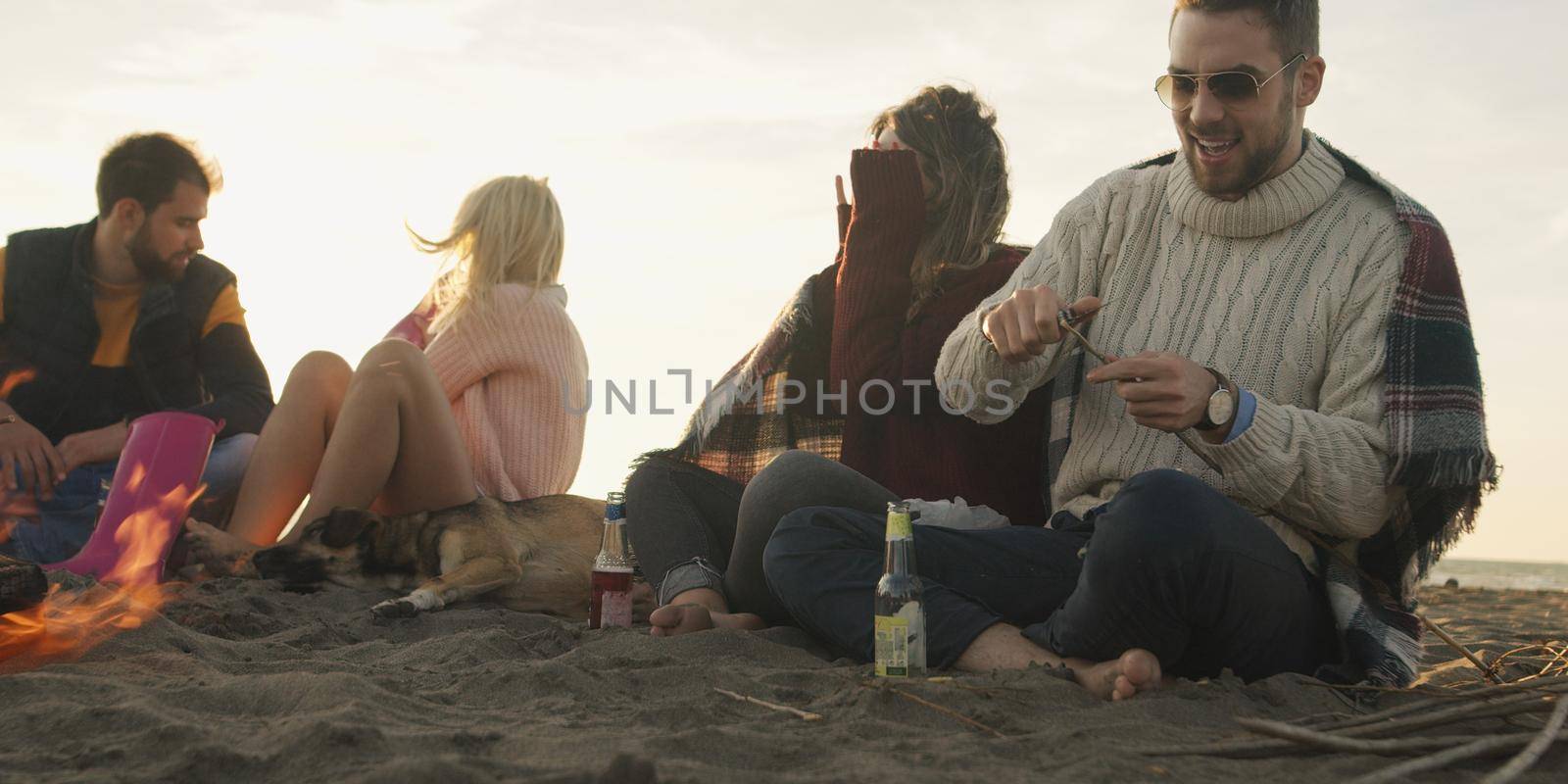 Group of friends with dog relaxing around bonfire on the beach at sunset