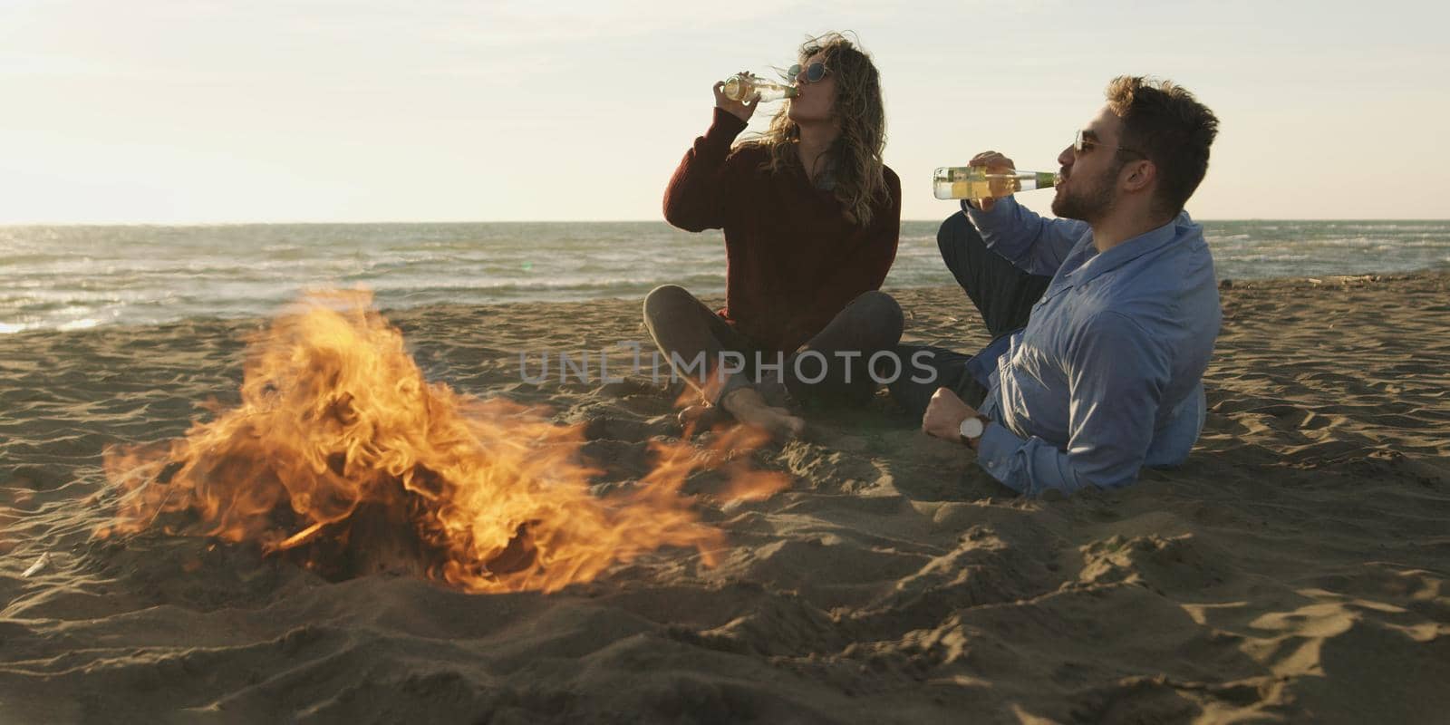 Loving Young Couple Sitting On The Beach beside Campfire drinking beer by dotshock