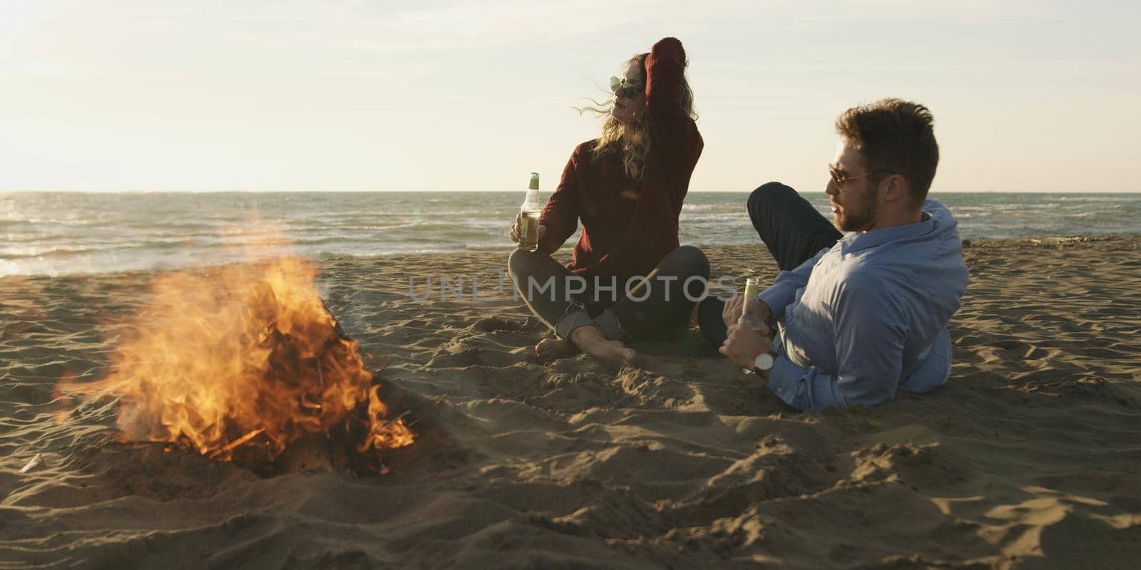 Young Couple Relaxing By The Fire, Drinking A Beer Or A Drink From The Bottle.