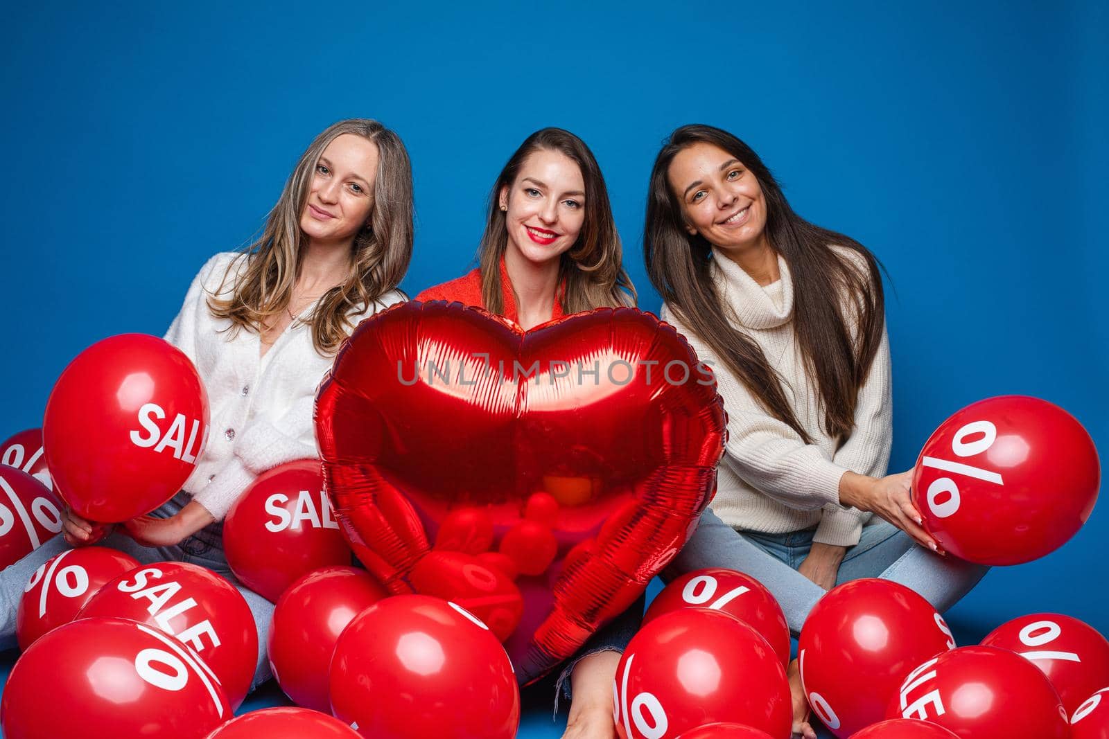 Portrait of three pretty smiling girlfriends in casual clothes sitting with red air balloons with sale and percent sign. Girl in the middle holding red heart-shaped balloon. Cutout on blue.