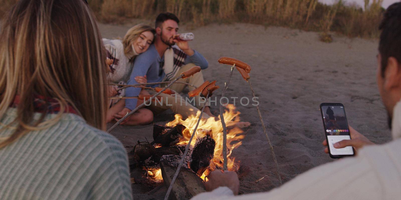 Group of young friends sitting by the fire late at night, grilling sausages and drinking beer, talking and having fun