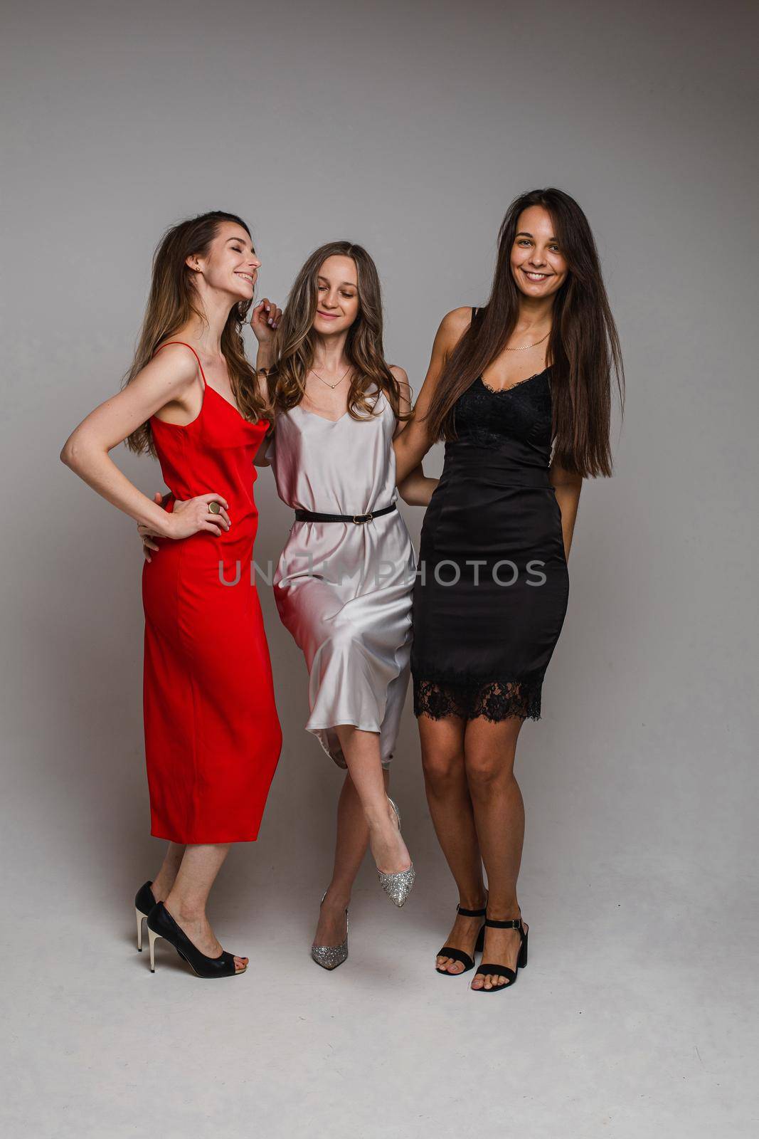 Charming positive young girls friends wearing stylish feminine evening dresses posing on gray studio background by StudioLucky