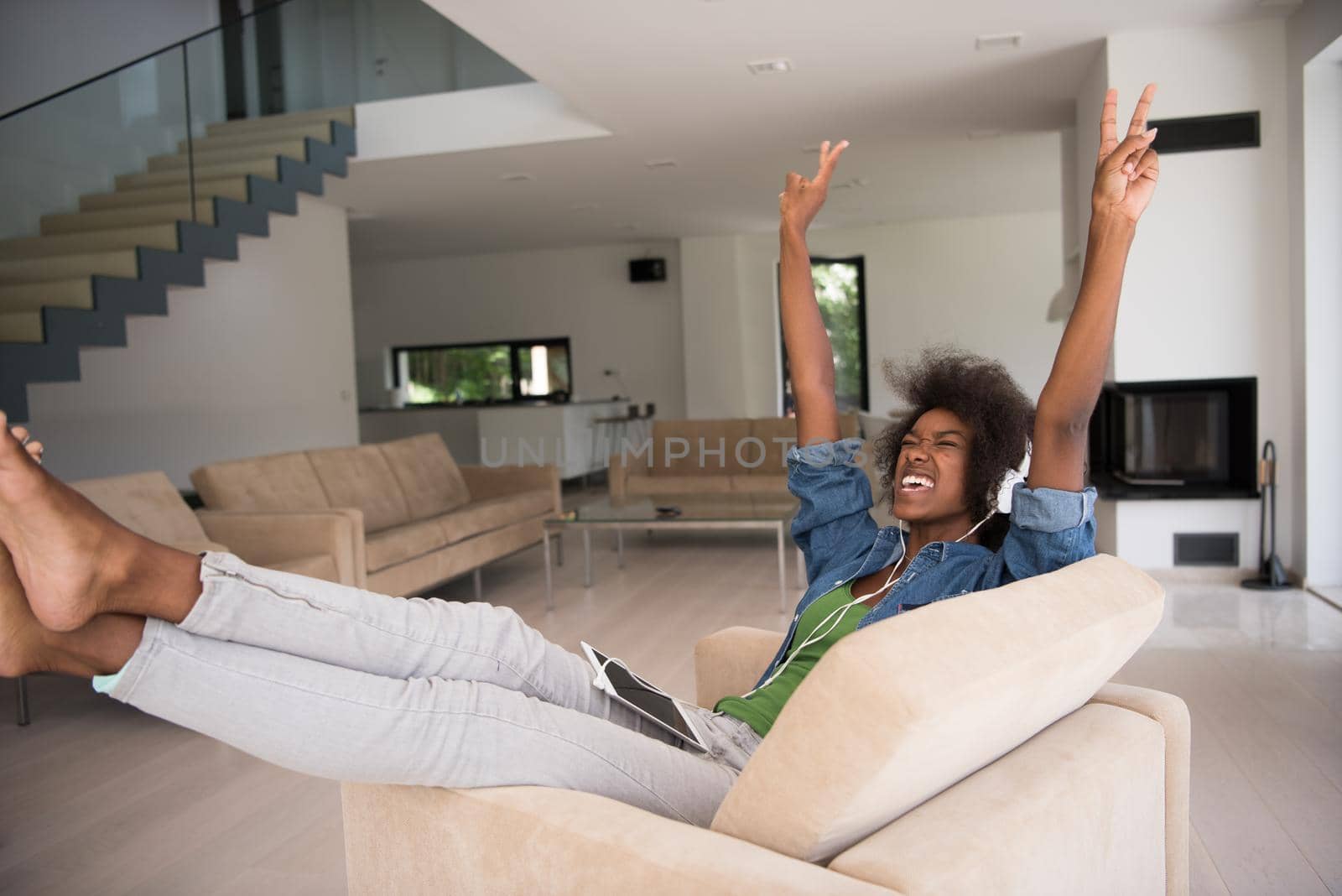 young beautiful African American woman enjoys listening to music with headphones and tablet in your armchair in her luxury home