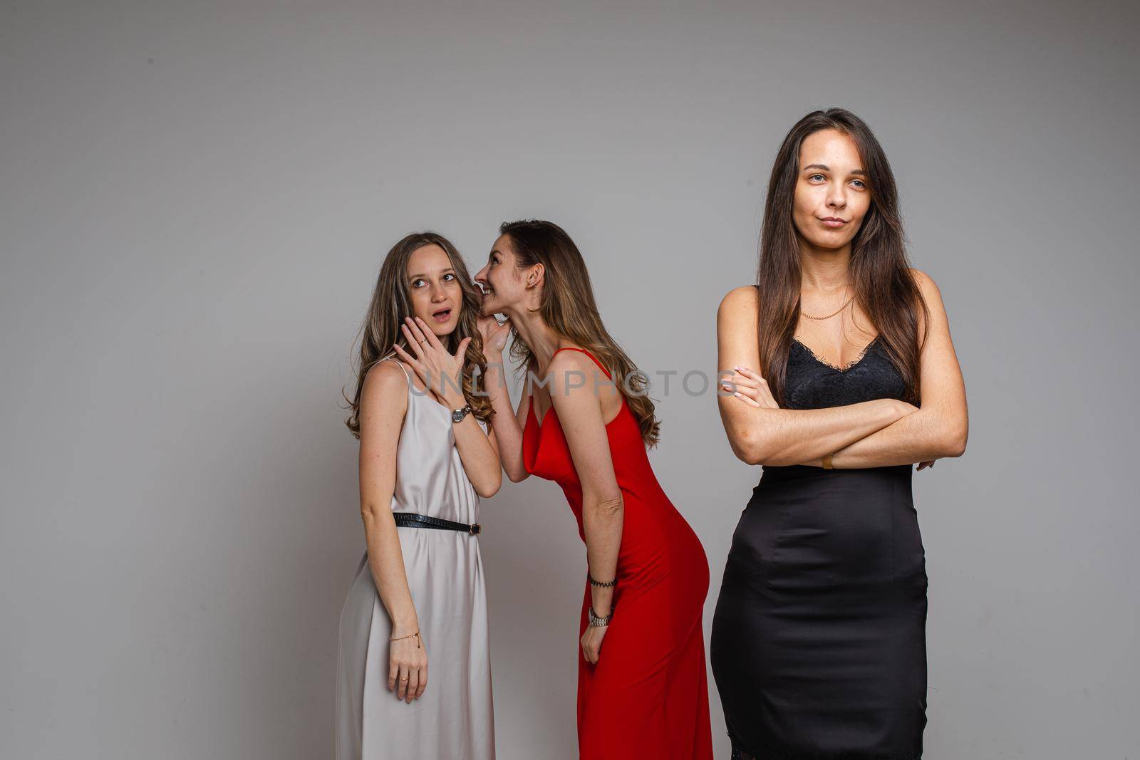 Studio portrait of gossiping girls in silver and black dresses mocking beautiful unhappy girl with long fair hair in red dress standing in the foreground and looking down sadly.